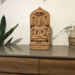 detailed, eye-level shot showcases an intricate sandstone carving of a crowned Bodhisattva, possibly Maitreya, seated in a meditative Dhyanasana posture on a multi-tiered lotus pedestal. The figure's hands are positioned in the Dhyana Mudra (meditation gesture), conveying deep serenity and contemplative focus. The Bodhisattva wears an elaborate crown, a long necklace, and armlets, with elongated earlobes typical of Buddhist iconography. The central figure is flanked by two smaller standing attendant figures, and the entire composition is set within a beautifully carved, arch-shaped stele. Above the Bodhisattva's head, a stylized Bodhi tree or similar foliage forms a canopy, with a parasol-like element indicating reverence. Celestial beings or devas holding offerings are carved into the upper corners of the stele, adding to the spiritual narrative. The base of the stele features a frieze with finely detailed relief carvings, possibly depicting mythological animals or auspicious symbols like rabbits and other creatures, further enriching the artifact's historical and cultural significance. The carving's earthy, reddish-brown sandstone texture highlights its ancient craftsmanship and the intricate details etched into the material. This spiritual artifact is prominently displayed indoors on a sleek, grey, reflective countertop, suggesting a modern yet culturally rich home decor aesthetic. A plain white wall serves as a minimalist backdrop, allowing the sculpture to stand out. To the left, a vibrant green houseplant with broad, pointed leaves (likely a Dracaena) adds a touch of natural life and contrast. On the right, a rustic, off-white ceramic vase, adorned with traditional patterns in warm tones, holds dried sprigs, complementing the organic feel of the sandstone. This image effectively captures the blend of ancient Buddhist art and contemporary interior styling, perfect for an article discussing spiritual home decor, the symbolism of sandstone sculptures, or cultural artifacts.