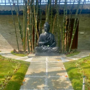 highly detailed, large dark grey Buddha statue sits serenely in the Dhyana Mudra (meditation) pose, positioned centrally on a sturdy matching stone plinth within a meticulously designed garden. The statue, featuring finely sculpted robes and a peaceful, contemplative countenance, anchors this tranquil outdoor space. Directly behind the Buddha, a dense cluster of towering, slender bamboo stalks provides a natural, vibrant green-brown screen, enhancing the calm atmosphere. In the background to the left, a modern building wall clad with varied shades of grey stacked stone is visible, suggesting a harmonious blend of natural materials and architectural design. A dark wooden door or panel is partially visible on the right side of the background. An inviting garden pathway stretches from the foreground directly towards the statue, comprised of large, flat light grey square stepping stones perfectly fitted together and separated by bright white decorative river pebbles. On either side of the path, lush, manicured green grass lawns are bordered by thin strips of white pebbles and delicate ornamental plants, contributing to the clean, zen-like aesthetic. The bright natural daylight illuminates the entire scene, emphasizing the textures of the various stone elements – from the smooth Buddha statue and stepping stones to the rougher stacked wall – and the vibrant greenery. This image beautifully illustrates the artistry of landscape design and the quality of stone garden decor, highlighting how natural stone products can be used to create peaceful and contemplative outdoor meditation spaces or zen gardens. The "The Stone Studio" watermark on the statue subtly indicates their craftsmanship and expertise in providing high-quality stone statues and landscaping solutions.