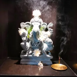 magnificent, hand-carved Ganesha statue, fashioned from dark grey natural stone with visible textural variations, dominates the center of a dimly lit, minimalist display. The revered Hindu deity, seated in a traditional posture, features an elephant head, large ears, and four arms. Its trunk is gracefully curved, and its upper left hand holds green foliage, while the right hand is raised in a gesture of blessing (Abhaya Mudra). The statue is adorned with delicate white daisy-like flowers – one prominently placed on its crown, others nestled around its neck and shoulders, and a single bloom resting at its base on the multi-tiered stone pedestal. A subtle "the Stone Studio" logo is etched into the lower front of the Ganesha idol, indicating its origin. To the right of the statue, two incense sticks burn steadily in a small, gleaming golden brass holder, releasing ethereal white smoke that curls elegantly upwards and drifts to the right, beautifully illuminated against the deep black background. This smoke creates a serene, almost mystical ambiance. On the statue's dark stone base, in front of Ganesha, sits a tiny, light-colored (likely white stone or ceramic) figure of Mushika, his traditional mouse mount. The entire scene is bathed in focused, dramatic lighting that highlights the intricate details of the carving and the flowing incense, emphasizing the spiritual and meditative atmosphere. This image likely connects to an article discussing artisanal stone craftsmanship, devotional sculptures, or the artistic creations of "the Stone Studio."