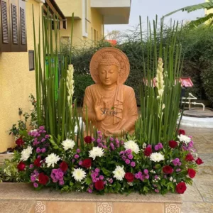 serene and intricately composed outdoor display centers around a medium-sized, light brown terracotta or sandstone Buddha statue, depicted seated in a meditative posture. The Buddha has a peaceful countenance with downcast eyes, a prominent ushnisha (cranial bump), and urna (dot between the eyebrows), and is draped in flowing robes. Its right hand is raised in a teaching or discussion mudra (possibly Vitarka Mudra), with the thumb and forefinger touching. Behind its head is a beautifully carved, circular halo or aura, detailed with repetitive patterns. The entire statue exudes a sense of tranquility and spiritual calm. The Buddha statue is the focal point of a vibrant and lush floral arrangement. On both sides, tall, slender green reeds or grasses rise vertically, framing the statue, and are interspersed with elegant spires of delicate white flowers, resembling tuberoses, adding height and texture to the display. Below, a dense and colorful bed of flowers stretches across the foreground, featuring numerous rich red roses, prominent clusters of white chrysanthemums (or similar daisy-like blooms), and vibrant splashes of smaller purple-magenta flowers, all nestled within dark green foliage. The entire arrangement rests on a decorative, light brown tiled platform, which features an ornate, symmetrical pattern on its vertical risers. In the background, to the left, a portion of a light yellow building with dark brown window shutters is visible, while to the right, a dense mass of green shrubbery and trees provides a natural, verdant backdrop, suggesting a peaceful garden or courtyard setting. The image, subtly watermarked with "the Stone" on the statue, beautifully captures themes of spiritual decor, tranquil garden design, handcrafted stone artistry, and the creation of serene meditation spaces, making it ideal for articles related to home garden enhancements, outdoor spiritual art, or landscape design.