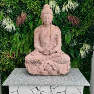 beautifully crafted Buddha statue, rendered in a striking reddish-pink speckled granite, sits serenely in the center of the frame. The statue is depicted in a traditional meditative posture (Padmasana or full lotus pose), with its hands resting gently in its lap in the Dhyana Mudra, symbolizing deep concentration and enlightenment. The natural stone texture is visible, showcasing subtle variations in color from rosy pink to deeper rust tones, giving it an earthy yet refined appearance. Intricate details are carved into the stone, including the soft folds of the Buddha's monastic robes, the elongated earlobes, the ushnisha (cranial bump signifying wisdom), and a peaceful, contemplative expression with eyes softly closed. The statue is positioned on a sturdy, rectangular pedestal. The top surface of the pedestal is a smooth, grey speckled granite, providing a clean contrast to the statue. The sides of the pedestal are faced with irregularly shaped white and light grey stone tiles, creating a textured, rustic mosaic effect that grounds the piece. Behind the entire arrangement is a vibrant and lush green vertical garden or living wall. This verdant backdrop is composed of various types of artificial foliage, including dense ferns and other leafy greens, interspersed with spiky white, light green, and reddish-brown ornamental plants or dried grasses, which add depth and visual interest. The overall scene exudes a profound sense of tranquility, peace, and natural beauty. A subtle watermark, "the Stone Studio," is visible on the lower left of the Buddha's lap, indicating the artisan or supplier of this elegant granite sculpture, which would be perfect for a zen garden, meditation space, or as a focal point in home and outdoor decor. This detailed alt text provides keywords for spiritual decor, stone art, garden sculptures, and tranquil outdoor living spaces.