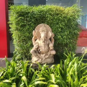 beautifully detailed, light-colored carved stone Ganesha statue is prominently displayed outdoors, set amidst vibrant greenery. The Hindu deity, Ganesha, with his iconic elephant head and four arms, is depicted seated in a serene posture. His upper right hand is raised in the Abhaya Mudra, offering blessings and fearlessness, while his lower left hand gently holds a Modak (sweet). Intricate carvings adorn his crown, jewelry, and the lotus petal base he sits upon, as well as the decorative arch or prabhamandala behind his head, which features a radiant, sun-like design. The texture of the stone appears smooth yet robust, highlighting the meticulous craftsmanship of this Ganesha idol. The statue is positioned centrally against a dense, lush backdrop of tall, vibrant green bamboo foliage, creating a striking natural contrast. In the foreground, lower-growing broad-leafed green plants, possibly including a white spider lily bloom on the right, add depth and a sense of a well-maintained garden or landscaped area. Hints of a modern building with large glass windows and a dark red panel are visible in the far background, suggesting an urban or commercial setting for this tranquil outdoor display. The natural sunlight illuminates the scene, emphasizing the peaceful ambiance. A subtle 'The Stone Factory' logo is visible on the lower left of the statue, indicating the source or artisan of this elegant stone sculpture, perfect for garden decor, spiritual spaces, or sophisticated landscaping projects showcasing fine stone artistry.