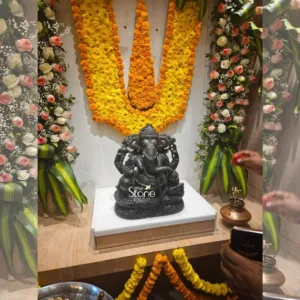 This image captures a beautifully arranged devotional setting, likely for an inauguration or a blessing ceremony, centered around an exquisitely crafted Ganesha idol. In the foreground, a majestic black stone statue of Lord Ganesha is prominently displayed on a clean white pedestal, which rests on a natural wood surface. The Ganesha idol is intricately carved, depicting the deity with multiple heads and arms, seated gracefully with remarkable detail on a textured base. The dark, polished stone highlights the expert craftsmanship, from the delicate features of his face and trunk to his elaborate crown and traditional attributes held in his hands. A watermark for "The Stone Studio" is subtly visible on the base of the idol, suggesting the origin or significance of this particular piece and its connection to the article's focus on stone craftsmanship or a related business. The background is adorned with vibrant floral decorations against a light-colored wall. A large, U-shaped arch of brilliant yellow and orange marigold garlands creates a festive and auspicious backdrop directly behind the Ganesha statue. Flanking this central marigold arch are two tall, vertical floral arrangements. These arrangements feature an elegant mix of delicate white and light pink roses, interspersed with tiny white baby's breath and long, slender green leaves, adding a touch of sophistication and freshness to the traditional setting. On the right side of the image, a hand with brown skin tone is visible, holding a small red flower petal, poised as if in the act of offering it during a puja or spiritual ritual. Below this hand, on the wooden surface, sits a traditional brass or copper pot, possibly a kalash, topped with a small golden figurine, further emphasizing the ceremonial atmosphere. In the lower right corner, another hand holds a modern smartphone, pointing it towards the display, suggesting that the event is being documented or shared, bridging traditional practices with contemporary engagement. More yellow and orange marigold garlands are visible along the bottom edge, indicating that the festive decorations extend beyond the immediate focal point. The overall scene conveys an atmosphere of reverence, celebration, and artistic appreciation, perfectly illustrating a blend of spiritual devotion and master craftsmanship, likely celebrating a significant milestone related to "The Stone Studio" or a showcase of their artisanal products.