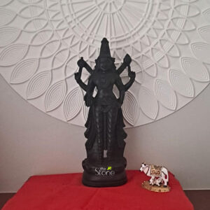 A serene interior arrangement showcasing an 18-inch Lord Vishnu statue hand-carved from natural black stone. The deity is depicted in a majestic standing posture atop a circular lotus pedestal positioned on an altar covered with a vibrant red silk cloth. The statue’s dark matte finish provides a striking contrast against a sophisticated white background wall, which features a large circular relief panel with a repetitive petal or fan-like design creating a divine halo effect around the idol. To the right of the Vishnu idol sits a small intricately decorated white cow figurine symbolizing the sacred Kamadhenu. The ambient lighting casts soft shadows, enhancing the spiritual and calming atmosphere of the space. This devotional setup serves as a prime example of an elegant spiritual nook for a home mandir illustrating the ideal placement of an 18-inch Vishnu statue to invite peace stability, and auspicious energy into a modern living environment.
