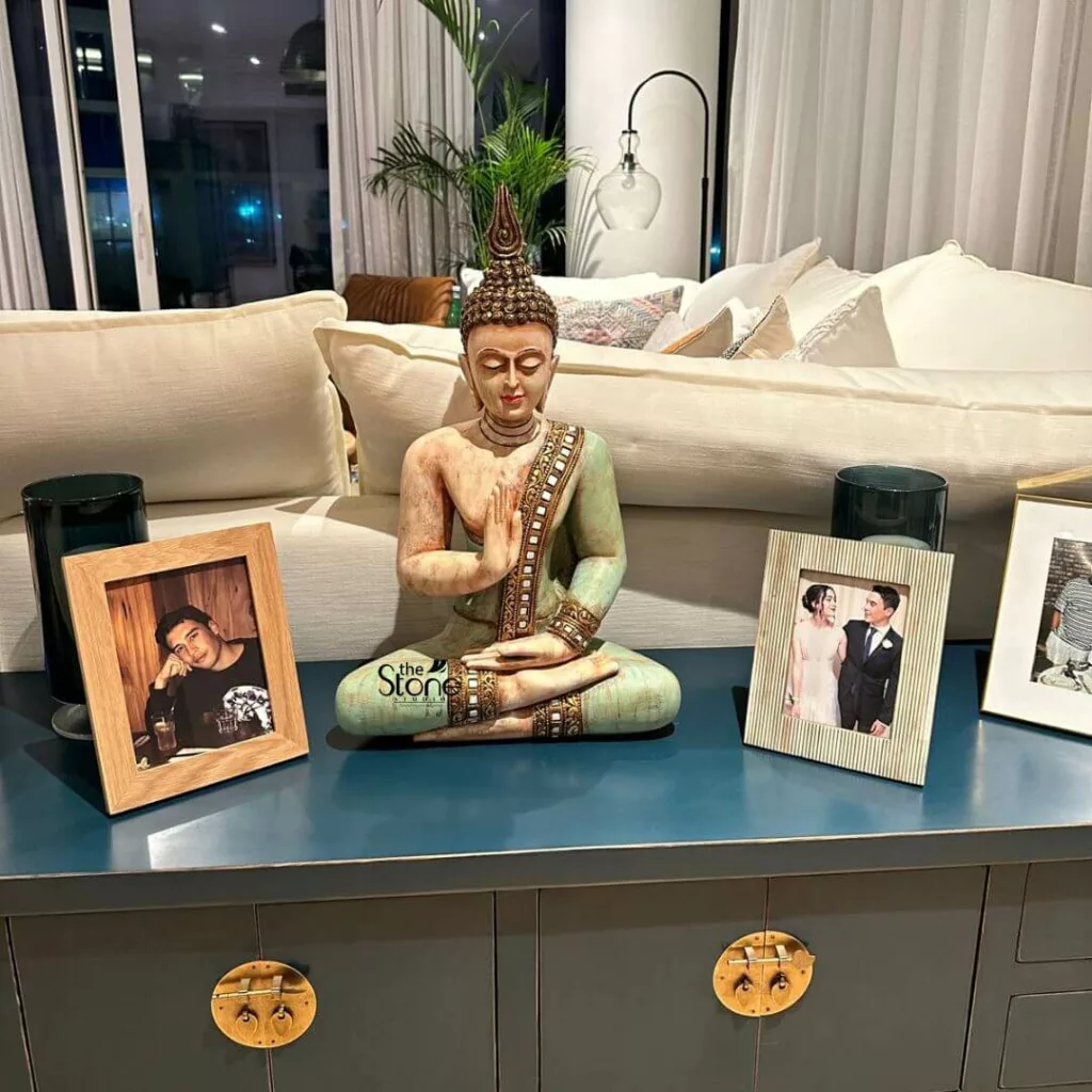 Decorating Apartment with small Statues 24 meticulously arranged and serene home decor display, centered on a dark teal-blue console table in a modern living room. Dominating the center is a beautifully detailed Buddha statue, depicted in a meditative lotus position (Padmasana) with hands gently pressed together in the Anjali Mudra (prayer or greeting) gesture. The statue features a light, natural stone-like complexion, adorned with serene jade-green robes that are intricately detailed with rich gold and dark brown patterns on the trim and sash. Its elaborate, textured golden headpiece (ushnisha) adds to its spiritual aesthetic, and its eyes are closed in peaceful contemplation. Flanking the Buddha statue are personal touches that add warmth and character to the space. To the left, a light natural wood photo frame holds a portrait of a young man with dark hair, resting his head on his hand and looking contemplatively at the viewer. On the right, a stylish rectangular photo frame with a distinctive cream and vertical striped pattern (possibly made of mother-of-pearl inlay or a similar textured material) showcases a joyful photograph of a couple, likely from a wedding or formal event, with the man in a suit and the woman in a white dress. A third, gold-framed photo is partially visible on the far right. Completing the symmetrical arrangement around the Buddha are two sleek, dark teal cylindrical vases or candle holders. The console table itself, a sophisticated dark blue or slate grey, features unique, ornate brass or gold-toned hardware on its cabinet doors, resembling stylized butterfly wings or traditional Asian lock mechanisms, hinting at an eclectic or globally inspired aesthetic. In the soft-focused background, a plush, inviting off-white or cream-colored sofa with numerous soft cushions creates a comfortable lounge area. Behind it, a large window reveals the gentle glow of distant city lights against a dark night sky, framed by elegant, sheer white curtains. A tall, leafy green houseplant, possibly a palm or similar tropical variety, adds a touch of natural vitality and freshness to the corner. Above the sofa, a modern black metal floor lamp with a clear glass globe shade arches gracefully, providing ambient lighting. This detailed image conveys a tranquil and thoughtfully curated living space, blending spiritual elements with cherished personal memories and contemporary design, suggesting a focus on mindfulness, personal sanctuary, or elegant home styling.