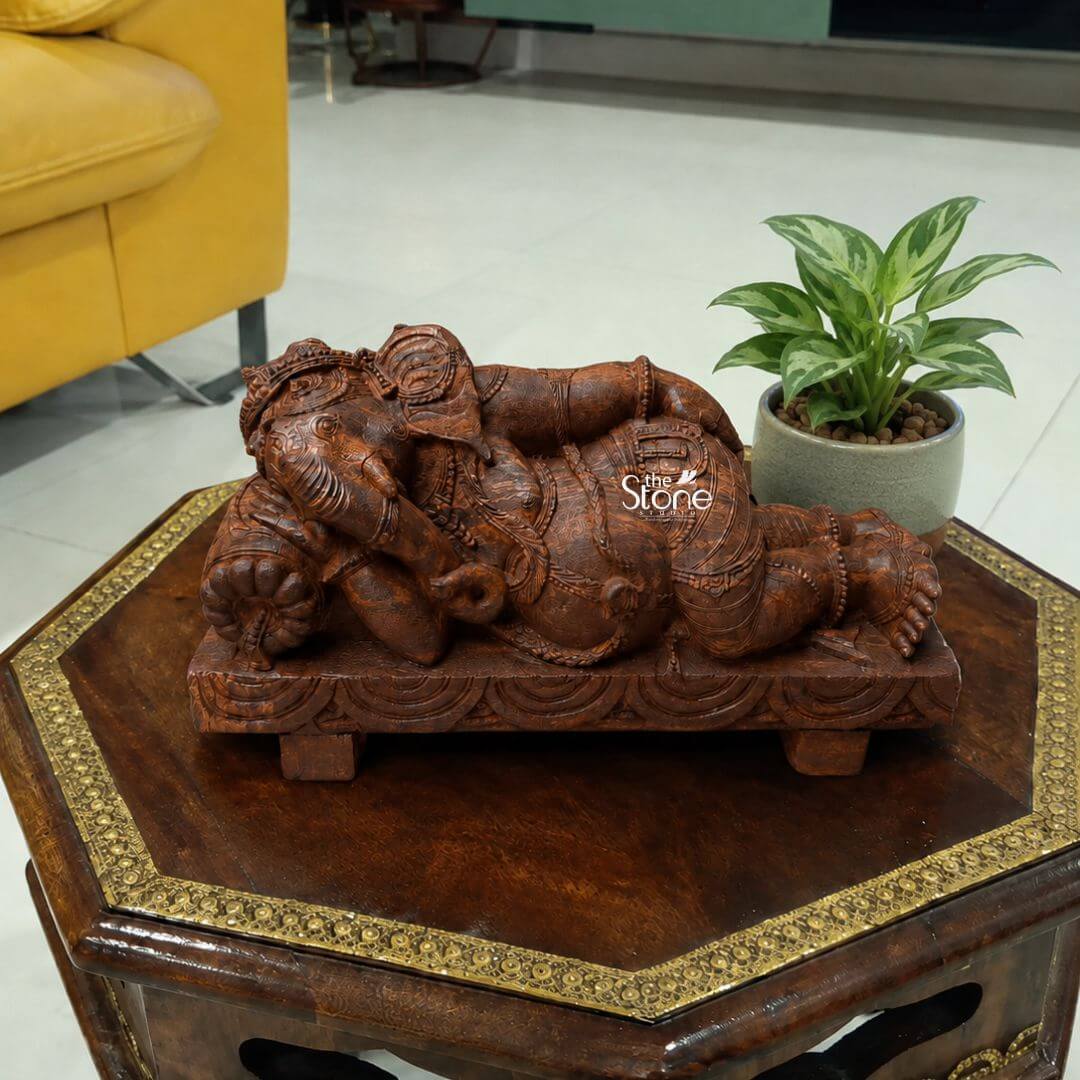 Sleeping Ganesh Murti 15" 3 A detailed medium shot captures a fifteen-inch-tall statue of Lord Ganesha meticulously hand-carved from a warm rich-brown sandstone. The statue depicts Ganesha in a graceful serene sleeping posture resting comfortably on his left side. His elephant head is supported by a small decorative pillow and he is lying on a matching ornately carved rectangular plinth with stylized feet. The deity is adorned with intricate shallow-relief carvings of elaborate jewelry a beaded crown and a flowing sacred thread showcasing the artisanal skill and the natural granular texture of the sandstone. The Sleeping Ganesha sculpture is positioned centrally on a dark octagonal wooden side table with a decorative hammered brass inlay pattern around its edges. The table surface provides a stable and traditional base contrasting beautifully with the light-colored stone. The background is a softly blurred modern interior living space featuring a bright mustard-yellow sofa to the left and a glimpse of a contemporary kitchen counter in the distance. This juxtaposition of traditional art with a modern setting highlights the statue's versatility as an elegant home decor piece. To the right of the Ganesha statue a potted indoor plant with variegated green and white leaves likely a type of Aglaonema adds a touch of vibrant natural life to the arrangement. The plant sits in a simple textured grey ceramic pot with small white pebbles on top. The lighting is soft ambient indoor light filtering from the side casting gentle shadows that emphasize the depth of the carving and the rich color of the sandstone. This harmonious composition creates a peaceful and inviting vignette perfectly illustrating the statue's suitability for enhancing Ludhiana homes.
