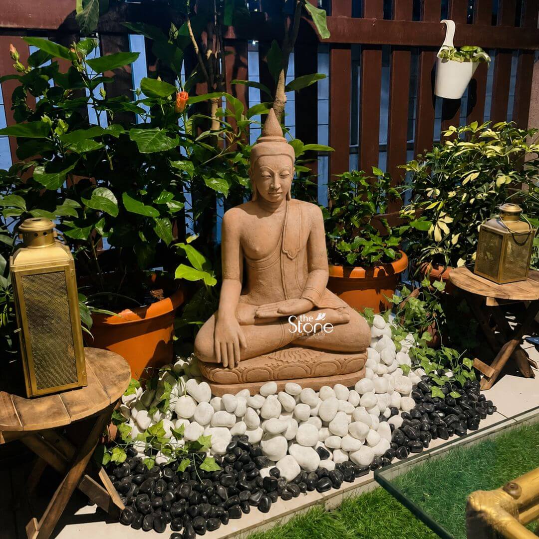 Bhumisparsha Buddha 3ft 3 A tranquil 3-foot Sandstone Buddha statue serving as an exquisite Buddha Stone Statue for Home Decor is beautifully integrated into a serene outdoor patio or balcony garden. The statue carved from warm reddish-brown sandstone depicts Buddha in a meditative seated posture specifically the full lotus position symbolizing deep concentration and spiritual enlightenment. His hands are in the Dhyana Mudra with both hands resting in his lap palms facing upwards and thumbs touching signifying meditation and inner peace. The natural earthy hue of the sandstone gives the sculpture an authentic calming presence blending seamlessly with its surroundings. The Buddha's facial features are finely carved radiating profound peace and wisdom with eyes gently closed or cast downwards in contemplation. His head is adorned with a conical topknot symbolizing his spiritual wisdom. The flowing drapery of his robes is sculpted with soft elegant folds emphasizing the texture of the sandstone and contributing to the statue's graceful and tranquil form. The statue is seated directly on a base of contrasting black and white river stones arranged artfully to create a striking visual pattern. This arrangement creates a dedicated zen-like space around the figure. The Buddha statue is surrounded by lush green potted plants of various sizes some with hints of red flowers creating a vibrant and natural backdrop against a dark wooden slatted fence. Two antique-style golden lanterns rest on small wooden side tables flanking the Buddha adding a warm inviting glow. In the foreground part of a glass-top table suggests a comfortable seating area. This 3-foot Sandstone Buddha statue is an ideal example of home decor designed to bring blessings peace and aesthetic beauty to any living space perfectly aligning with an article focusing on such serene and contemplative Buddha stone statues.