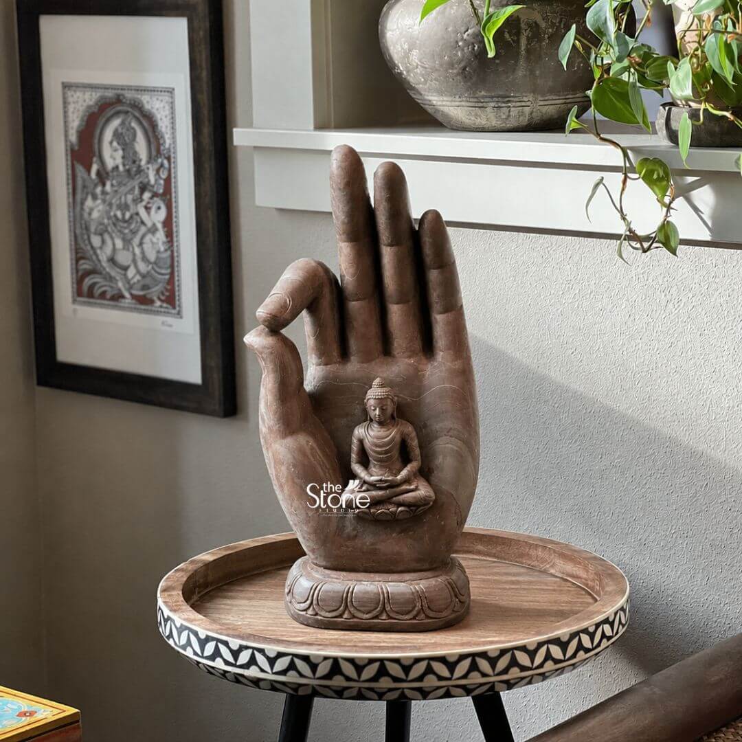 Buddha Hand Statue 16" 3 A unique 16-inch Hand Buddha Statue carved from pink stone serves as a prominent and artistic decorative element on a stylish round wooden table enhancing a cozy home interior. The sculpture depicts an intricately detailed human hand palm facing upwards with a small serene Buddha figure seated within its grasp. The hand itself forms a symbolic mudra possibly the Vitarka Mudra with the thumb and forefinger touching symbolizing wisdom and discourse. The pink stone which has a warm earthy tone gives the entire sculpture a natural and calming aesthetic. The small Buddha figure seated in the palm is depicted in a meditative posture with downcast eyes and a peaceful expression mirroring the larger theme of tranquility. The base of the hand sculpture features carved lotus petal designs adding a traditional touch. In the background a framed piece of artwork depicting a deity hangs on the wall and a shelf holds a potted plant contributing to the overall harmonious decor. This 16-inch pink stone Buddha Hand statue is an excellent visual for an article about Buddha hand statues for home decor. Its unique design combining a symbolic hand gesture with the serene Buddha makes it a powerful focal point for meditation or simply as a piece that radiates peace and wisdom. The warm tones and artistic craftsmanship make it a versatile addition to various interior styles perfect for bringing a sense of spiritual calm and aesthetic beauty to any living space.