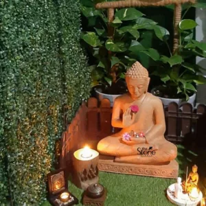 meticulously arranged serene meditation corner or indoor zen garden, showcasing a prominent, handcrafted sandstone Buddha statue as its focal point. The medium-sized Buddha, rendered in a warm, earthen-toned sandstone, sits gracefully in the Dhyana Mudra, a meditative lotus position, upon an intricately carved pedestal featuring swirling patterns. Its right hand gently cradles a vibrant red rose, while the left hand holds a small offering of delicate pink and purple flowers in a tiny bowl, symbolizing compassion and enlightenment. The Buddha's face wears a serene, tranquil expression with softly closed or downcast eyes, and a distinct ushnisha (topknot) crowns its head, signifying wisdom. The spiritual display is set on a lush, vibrant green faux grass mat, creating the illusion of a miniature natural landscape. To the left, a charming miniature rustic wooden picket fence acts as a boundary against a dense vertical garden of artificial small-leafed green foliage, resembling a meticulously trimmed boxwood hedge. On the right, a large, healthy Pothos plant with variegated green and yellow leaves thrives in a grey pot, contributing to the natural ambiance. A rustic, woven brown structure, possibly part of a trellis or hanging decor, is visible in the upper background. Numerous elements enhance the peaceful atmosphere and invite contemplation. To the Buddha's left, a glowing candle is set within a rustic, carved wooden log-slice holder, casting a soft, warm light. Below it, a dark-framed miniature artwork depicting a Buddha head reflects the light from another small flickering tealight candle. An elegant, dark bronze incense burner with an ornate lid rests on a small wooden stand nearby, ready for aromatherapy and spiritual practice. To the Buddha's right, a small, circular white tiered altar holds a collection of miniature spiritual figurines: a golden meditating Buddha, a white robed figure, and slender incense sticks, signifying offerings and devotion. The overall lighting is soft, warm, and inviting, primarily originating from the candles, creating a deeply tranquil and meditative ambiance. A watermark for 'The Stone Studio' is subtly visible on the Buddha statue, indicating the artisanal craftsmanship and potential source of these beautiful spiritual decor items, ideal for creating a peaceful sanctuary in any home or garden. This scene perfectly encapsulates the essence of mindfulness, relaxation, and creating a sacred space for meditation and spiritual well-being.