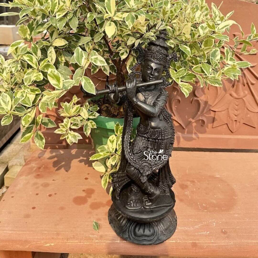 Black Stone Krishna Murti 21" 3 A beautiful 21-inch Black Stone Krishna statue perfectly sized as a Small Krishna Statue for Home is gracefully displayed on a warm terracotta-colored surface possibly a bench or shelf against a backdrop of vibrant variegated green and white foliage. The statue depicts Lord Krishna in his iconic Tribhanga pose with a gentle sway to his body radiating charm and divine playfulness. Carved from lustrous black stone the figure is exquisitely detailed with an elaborate crown intricate jewelry adorning his neck arms and ankles and finely sculpted garments. He holds a flute to his lips poised as if playing a melodious tune. The craftsmanship is evident in the delicate features of his face including his serene expression and the intricate detailing of his crown. The flowing drapery of his garments is intricately sculpted to show texture and movement clinging gracefully to his form. The black stone's smooth finish highlights these fine details creating a striking contrast with the lighter backdrop. The statue stands on a tiered round base adorned with lotus petal carvings adding to its spiritual and aesthetic appeal. The surrounding elements enhance its suitability as a home decor piece. A large potted plant with striking green and white leaves creates a lush natural backdrop while a carved terracotta wall or panel is visible behind the plant adding an earthy texture to the scene. This 21-inch Black Stone Krishna statue is an exquisite example of religious artistry designed to bring blessings peace and aesthetic beauty to any home making it an ideal focal point for devotion or decorative accent aligning perfectly with an article focusing on small Krishna statues for home interiors.