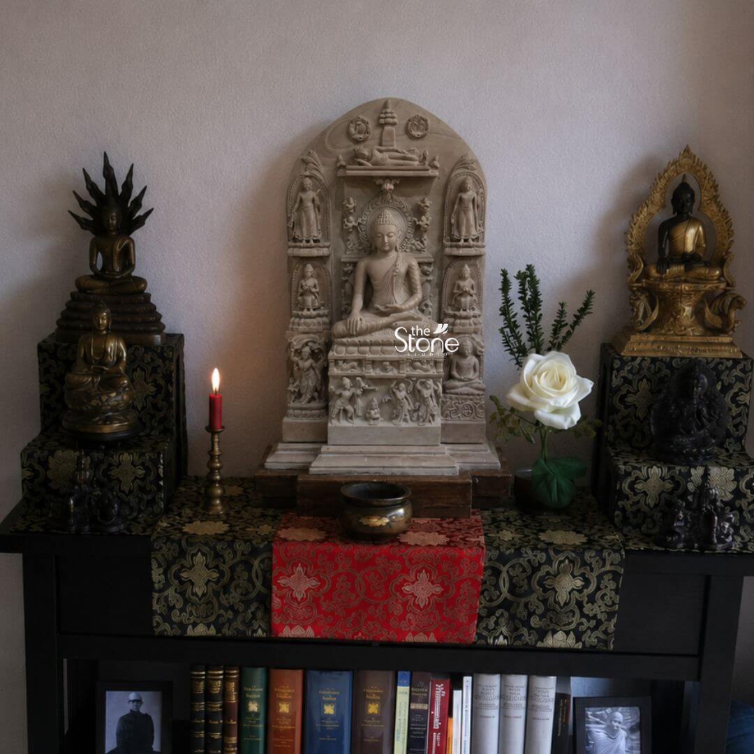 A beautiful 15-inch Buddha Soapstone statue is the central focus of an intricately arranged home decor display creating a sacred and meditative altar on top of a dark wooden bookshelf. The central Buddha is depicted in a traditional meditative seated posture with his hands resting in his lap emanating profound peace and spiritual wisdom. The soapstone gives the statue a soft earthy tone complementing the rich textures of the setup. The central Buddha statue is framed by an elaborate multi-tiered carved stone archway adorned with smaller figures and decorative motifs suggesting a temple-like structure. The details on his hair robes, and serene face are clearly visible. In front of the central Buddha a small brass singing bowl and a single white rose in a vase are placed on a wooden riser which is covered by a richly patterned red and gold fabric runner. To the left and right on separate dark pedestals draped with matching black and gold patterned fabric are two other Buddha figures: a bronzed Buddha with multiple serpent heads on the left and a golden Buddha in a decorative arch on the right further enhancing the spiritual ambiance. This 15-inch soapstone Buddha statue is an ideal subject for an article on Buddha statue decor for homes. The entire arrangement with its rich fabrics warm candle glow and variety of Buddha forms creates a harmonious and sacred space. The lit red taper candle in a brass holder to the left and the well-organized books in the shelves below suggest a blend of spiritual devotion and intellectual pursuit. This display offers a powerful example of how Buddha statues can be integrated into home decor to foster peace mindfulness and a sense of enlightenment.