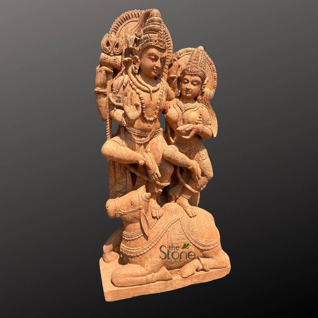 Shiv Parvati Idol Online 4ft 2 magnificent, intricately carved reddish-brown sandstone sculpture depicts the Hindu deities Lord Shiva and Goddess Parvati, accompanied by Shiva's divine vehicle, Nandi the bull. The central figure on the left is Lord Shiva, shown in a seated or relaxed posture with his left leg bent and resting comfortably. He is adorned with an elaborate crown (mukuta), multiple necklaces, armlets, and other traditional Hindu ornaments, with fine details highlighting his sacred thread (yajnopavita). His serene facial expression conveys divine wisdom and tranquility. His left hand is raised in a graceful gesture, possibly a blessing (abhaya mudra), while his right hand rests gently on his leg. Behind Shiva's head, an ornate, semicircular halo (prabhamandala) is intricately carved with flame-like or floral motifs, enhancing his divine aura. To Shiva's right, slightly lower and leaning in, stands Goddess Parvati, his consort. She is depicted with equal grace, wearing a detailed crown, necklaces, and a beautifully draped garment (sari or dhoti) with delicate pleats and patterns. Her hands are held together near her chest, possibly holding a small offering like a flower or a bowl, signifying devotion. Her expression is gentle and loving, complementing Shiva's composure. A smaller, matching halo frames her head, completing her divine depiction. At the base of the sculpture, Nandi the bull, Shiva's revered mount, is shown in a recumbent (lying down) position. Nandi is meticulously carved with decorative saddle-cloth patterns and traditional ornaments, looking upwards towards the divine couple with an expression of unwavering loyalty and devotion. The entire sculpture is crafted from a warm, reddish-brown sandstone, showcasing exceptional detail and artistry in traditional Indian stone carving. The rich texture of the material adds depth and character to the figures and their elaborate adornments. The deities stand on a simple, rectangular plinth. The plain, dark gray background highlights the sculpture, drawing full attention to its intricate craftsmanship and spiritual essence. A discreet watermark, "the Stone Studio," is visible in the bottom right corner. This sculpture, representing divine harmony and the sacred union of Shiva and Parvati (often referred to as Uma-Maheshwara), is a prime example of Hindu religious iconography and traditional Indian art, suitable for discussions on cultural heritage, spiritual artifacts, or elaborate stone craftsmanship.