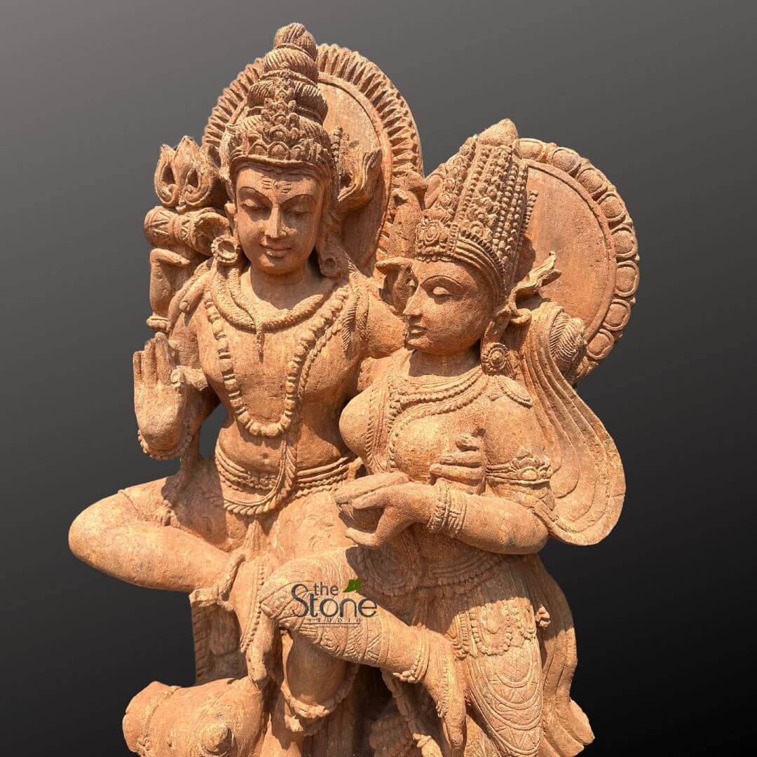 Shiv Parvati Idol Online 4ft 3 finely carved, large, light reddish-brown sandstone sculpture depicts the Hindu divine couple, Lord Shiva and Goddess Parvati, seated together in a posture of conjugal harmony and divine grace. The male figure, Shiva, on the left, is shown with an elaborate topknot crown (jata mukuta), serene facial features, and multiple necklaces, bracelets, and a sacred thread (yajnopavita). His right hand is raised in the Abhaya Mudra, symbolizing fearlessness and reassurance, while his left arm gently embraces Parvati. Behind him is a richly detailed circular aureole (prabhavali) with radiating, flame-like patterns. The female figure, Goddess Parvati, on the right, is depicted with a magnificent, multi-tiered crown (kirita-makuta) and elegant, flowing hair that cascades down her back. Her face is soft and graceful, gazing towards Shiva, conveying devotion and companionship. She is adorned with intricate jewelry including large earrings, multiple necklaces, bangles, armlets, and a detailed waistband (kamarbandh), her garments delicately carved to show folds and patterns. Her right hand appears to hold a small, possibly symbolic object, like a lotus bud or seed, in her lap. Both deities are rendered with classical Indian sculptural aesthetics, emphasizing their divine forms and elaborate traditional adornments. The sculpture exhibits a visible granular texture characteristic of sandstone, highlighting the artisan's skill in traditional stone carving techniques. The warm, earthy tones of the stone provide a striking contrast against a plain, dark grey background, drawing full attention to the exquisite details of this devotional Hindu idol. A watermark for 'the Stone Studio' is subtly visible on the lower front part of the sculpture, indicating the source of this exquisite piece of religious art or spiritual decor, suggesting its connection to craftsmanship, antique reproductions, or sales of stone deity sculptures.