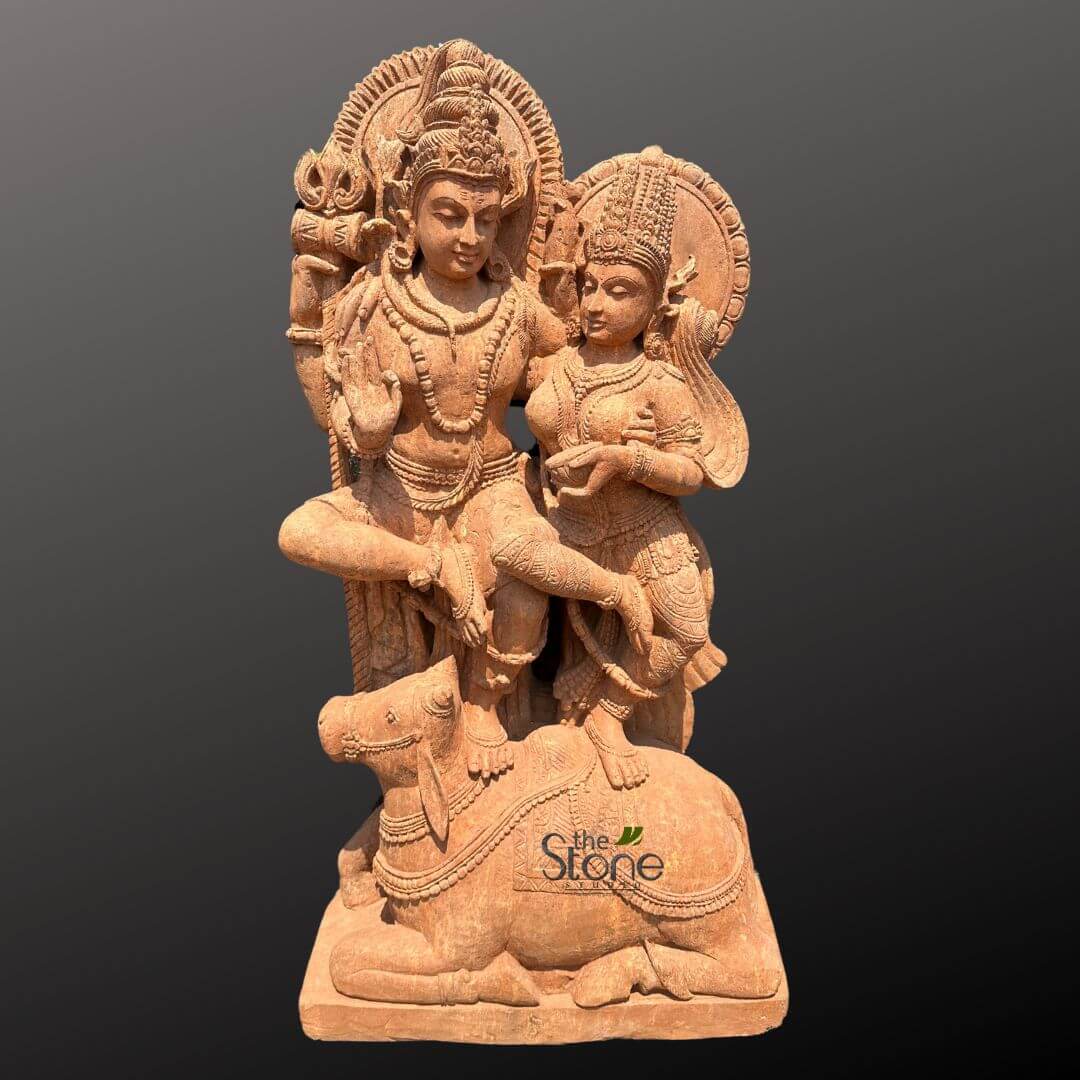 Shiv Parvati Idol Online 4ft 1 stunning, intricately carved reddish-brown sandstone sculpture depicts the revered Hindu divine couple, Lord Shiva and Goddess Parvati, in a moment of serene togetherness, seated gracefully upon Shiva's sacred bull, Nandi. The entire artwork, rendered in a warm, earthy terracotta-hued stone, showcases meticulous detail and traditional Indian craftsmanship. Lord Shiva is positioned on the left, seated in a dignified cross-legged posture atop Nandi's back. He is recognized by his elaborate tall crown (jatamukuta), serene facial expression with eyes gently cast downwards, and his right hand raised in the iconic Abhaya Mudra, a gesture of fearlessness and reassurance. A subtle trident (Trishula), his principal weapon, is visible emerging from behind his left shoulder, adding to his divine iconography. He wears multiple strands of beaded necklaces, armlets, and a sacred thread (yajnopavita), all intricately carved, showcasing a rich texture. A beautifully detailed, radiating circular halo (prabhamandala) frames his head. Goddess Parvati sits gracefully to Shiva's right, leaning gently towards him, symbolizing their eternal union. Her right leg is bent and rests on Nandi, while her left leg is also drawn in. She is adorned with a delicate, ornate crown, intricate earrings, necklaces, bracelets, and an elegantly draped sari or garment that accentuates her feminine form. Her hands are delicately posed, possibly holding a small offering or in a gesture of devotion, with a gentle, contemplative gaze complementing Shiva's meditative demeanor. A similar, though slightly smaller, halo frames her head, emphasizing her divine status. Beneath them, Shiva's faithful vahana (mount), Nandi the bull, lies recumbent on a simple rectangular plinth, forming a stable base for the divine pair. Nandi is richly decorated with beaded garlands, a saddle-cloth, and bells, emphasizing its sacred role and the reverence for all creation in Hindu philosophy. The entire sculpture exhibits remarkable craftsmanship, with every piece of jewelry, garment fold, and anatomical detail meticulously rendered, highlighting the skill of traditional Indian stone carving. The warm, earthy tones of the natural stone contrast subtly with the plain, dark gray background, allowing the spiritual aura, intricate details, and artistic mastery of the piece to truly stand out. This exquisite artwork, bearing a discrete "the Stone" logo on Nandi's side, serves as a powerful representation of devotion, cosmic balance, and the rich iconography found in Hindu temples and homes, making it an ideal piece for spiritual reflection or as a centerpiece of traditional Indian decor.