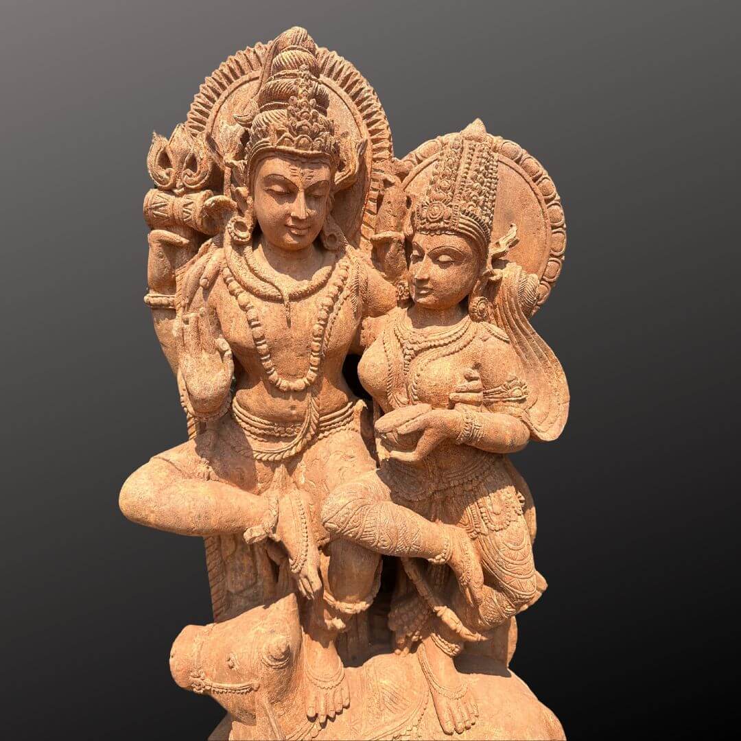 Shiv Parvati Idol Online 4ft 5 highly detailed, reddish-brown sandstone sculpture depicts the Hindu divine couple, Lord Shiva and Goddess Parvati, presented against a stark, dark grey background that accentuates its intricate forms. The artwork showcases Shiva, on the left, and Parvati, on the right, in a loving, seated embrace, symbolizing cosmic union and divine love. Lord Shiva is portrayed with a serene, contemplative expression, eyes slightly downcast. He wears an elaborate tiered crown adorned with detailed carvings, along with multiple necklaces, armbands, and a sacred thread. His muscular physique is subtly rendered beneath his dhoti-like garment. His right hand is raised in the Abhaya Mudra, a gesture signifying fearlessness and blessing, gently holding a lotus flower or similar sacred bloom. His left hand rests gracefully on his thigh. Shiva is seated with his right leg bent and raised, while his left leg rests upon the back of his sacred bull mount, Nandi, whose head is visible and facing left at the base of the sculpture. Behind Shiva's head is a circular halo, intricately carved with radial patterns that suggest divine light or a sun motif. Goddess Parvati is seated to Shiva's right, her body slightly turned towards him, reflecting devotion and companionship. She also wears an ornate crown, perhaps slightly different in style from Shiva's, along with delicate necklaces, earrings, armlets, and bangles. Her garment, a beautifully draped saree or skirt, is rich in texture and detailed pleats. Her hands are held forward in front of her, gently cradling a small vessel, bowl, or offering cup, a gesture of reverence or offering. A similar circular, radially patterned halo frames her head, emphasizing her divine status. The sculpture's earthy, reddish-brown hue and the finely chiseled details in the deities' attire, jewelry, facial features, and halos highlight the exceptional craftsmanship characteristic of traditional Indian religious art. This ancient style sculpture, likely made of sandstone or a similar material, serves as a powerful visual representation of Hindu mythology, devotion, and the profound spiritual significance of the divine feminine and masculine principles.