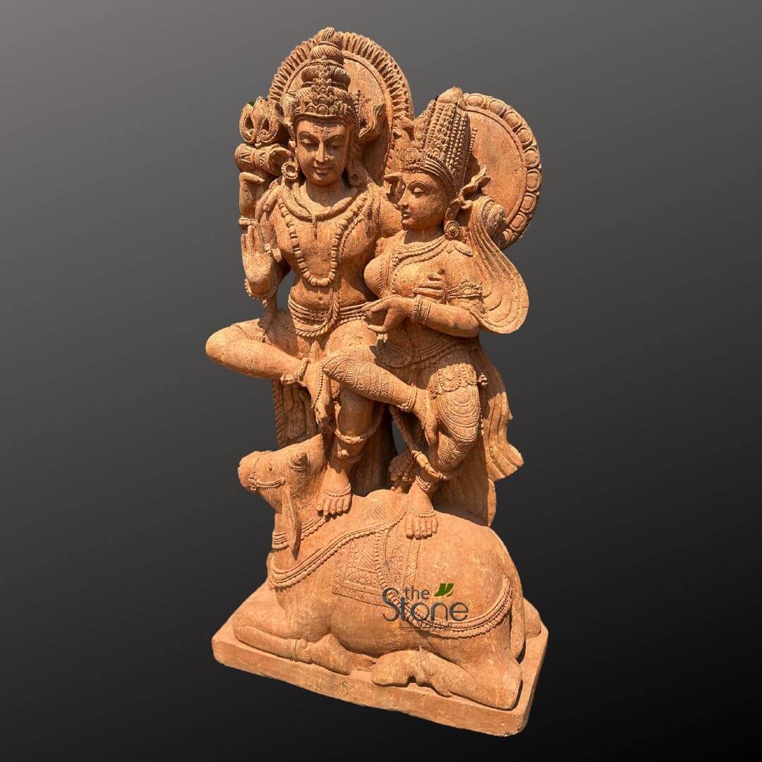 Shiv Parvati Idol Online 4ft 6 richly detailed reddish-brown stone sculpture depicts the Hindu divine couple, Shiva and Parvati (also known as Uma-Maheshwar), seated gracefully upon Shiva's sacred bull, Nandi. The entire sculpture is presented against a plain, dark gray background, allowing its intricate craftsmanship and devotional artistry to stand out prominently. Lord Shiva, positioned on the viewer's left, is depicted with a serene, compassionate expression, his left hand raised in the Abhaya Mudra, a gesture of fearlessness and protection, while his right arm is gently around Parvati. He wears an elaborate, tall crown (possibly Jatamukuta, signifying matted hair), multiple necklaces, armbands, bangles, and a meticulously draped lower garment, all carved with fine, traditional patterns. A subtle third eye may be discernable on his forehead. To his right, nestled affectionately on his left lap or beside him, sits Goddess Parvati, equally resplendent. She wears a towering, ornate crown, numerous pieces of finely carved jewelry including large earrings, multi-strand necklaces, armbands, bangles, and a gracefully flowing sari with intricate decorative motifs. Her right arm is subtly positioned around Shiva, while her left hand appears to hold a small, delicate object or make a gentle gesture near her lap. Her gaze is soft and contemplative, directed slightly downward or towards Shiva. Both deities are backed by beautifully carved, circular aureoles or halos with radiating patterns, emphasizing their divine status. Their facial features convey tranquility, grace, and eternal affection. Below them, Nandi, Shiva's sacred bull and mount, is carved in a reclining position, facing forward, with its head slightly raised in reverence. Nandi is adorned with a decorative saddle cloth featuring intricate patterns, along with necklaces and bells, symbolizing devotion, strength, and purity. The entire multi-figure composition rests on a simple, rectangular stone plinth, giving the impression of a substantial and stable piece of religious artwork. The terracotta-like reddish-brown hue of the sandstone or similar natural stone highlights the meticulous traditional Indian artistry and exceptional stone carving techniques used to create this piece. It reflects ancient Hindu iconography and the celebration of the divine couple, making it a significant representation of cultural heritage and spiritual devotion.
