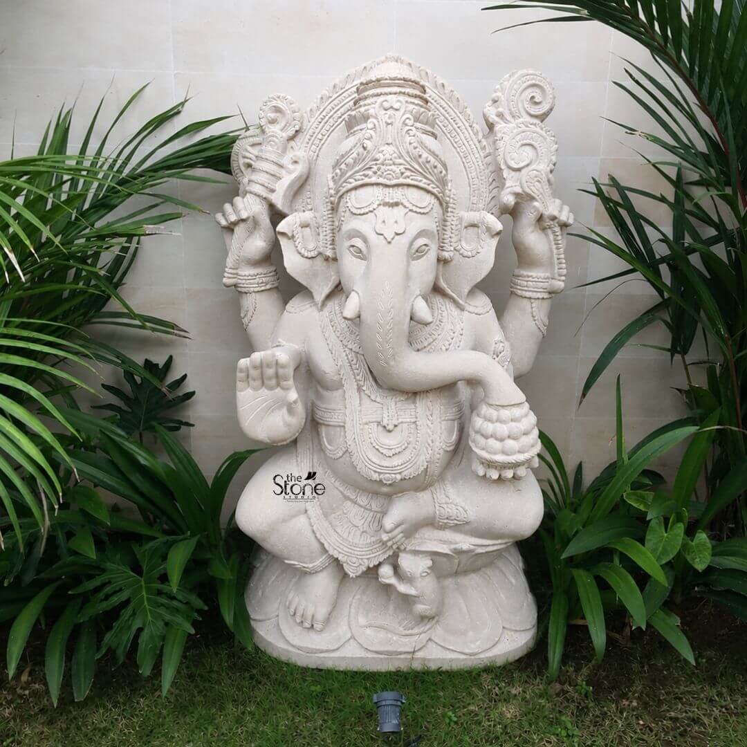 3 feet Ganesh Idol 3 A magnificent 3-foot Off-white Sandstone Ganesha statue perfectly suited as a Ganapathi Idol for a Garden stands majestically amidst lush green foliage against a light-colored textured wall. The statue depicts Lord Ganesha in a seated posture radiating an aura of wisdom and benevolence. His elephant head is finely sculpted complete with large ears expressive eyes and his iconic broken tusk. He is adorned with intricate traditional jewelry and a crown all carved from the same light sandstone which gives the sculpture a timeless and serene appearance. Ganesha is portrayed with four arms holding his customary attributes a modak in one hand an axe or goad in another and his lower right hand extended in a gesture of blessing offering reassurance and protection. The details on his garments the sacred thread and the patterns on his halo or Prabhavali behind his head are meticulously rendered showcasing the artistry of the carving. The off-white sandstone material naturally highlights these textures and details lending a soft earthy elegance to the deity. The statue is positioned on a neatly manicured green lawn framed by vibrant green plants with broad leaves on either side which grow against the light-colored wall. These plants create a natural and harmonious backdrop enhancing the spiritual tranquility of the idol and making it a focal point in any garden setting. This 3-foot Off-white Sandstone Ganesha statue is an exquisite example of a garden deity designed to bring blessings peace and aesthetic beauty to an outdoor space aligning perfectly with the article's focus on Ganapathi Idols for gardens.