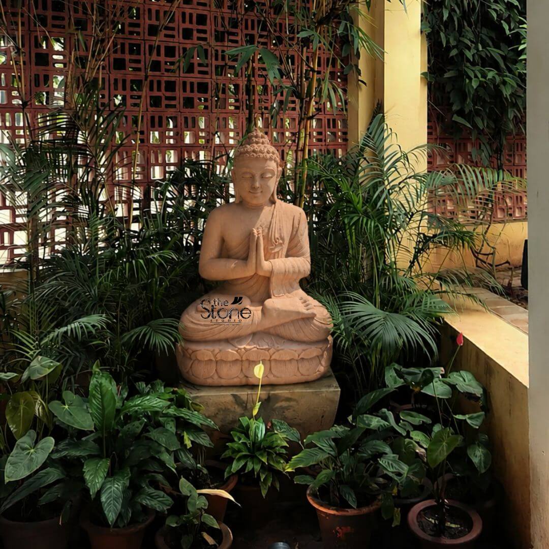 3 feet Anjali Mudra Buddha 3 A serene 3-foot Brown Sandstone Buddha statue serving as a tranquil Namaskar Buddha Idol for Garden is beautifully integrated into a lush and peaceful outdoor space. The statue carved from warm reddish-brown sandstone depicts Buddha in a meditative seated posture specifically the full lotus position symbolizing deep concentration and spiritual enlightenment. His hands are held together at chest level in the Anjali Mudra also known as Namaskar Mudra with palms pressed together and fingers pointing upwards signifying respect reverence and greeting. The natural earthy hue of the sandstone gives the sculpture an authentic ancient feel blending seamlessly with its organic surroundings. The Buddha's facial features are exquisitely carved radiating profound peace and wisdom with eyes gently closed or cast downwards in deep contemplation. His head is adorned with tightly coiled hair culminating in the ushnisha the cranial bump symbolizing his supreme spiritual knowledge. The flowing drapery of his robes is sculpted with soft elegant folds emphasizing the texture of the sandstone and contributing to the statue's graceful and tranquil form. The statue rests on a beautifully sculpted lotus-petal base which in turn sits on a simple square stone pedestal elevating the deity within its garden sanctuary. The Buddha is surrounded by a variety of lush green plants in pots and garden beds including palm-like foliage and smaller leafy plants creating a harmonious and serene environment. In the background a decorative terracotta brick lattice wall and tall bamboo shoots add texture and a sense of enclosure while a yellow pillar on the right suggests a nearby structure. This 3-foot Brown Sandstone Namaskar Buddha statue is an exquisite example of a garden deity designed to bring blessings peace and aesthetic beauty to any outdoor space perfectly aligning with an article focusing on such a revered and welcoming Buddha Idol for gardens.