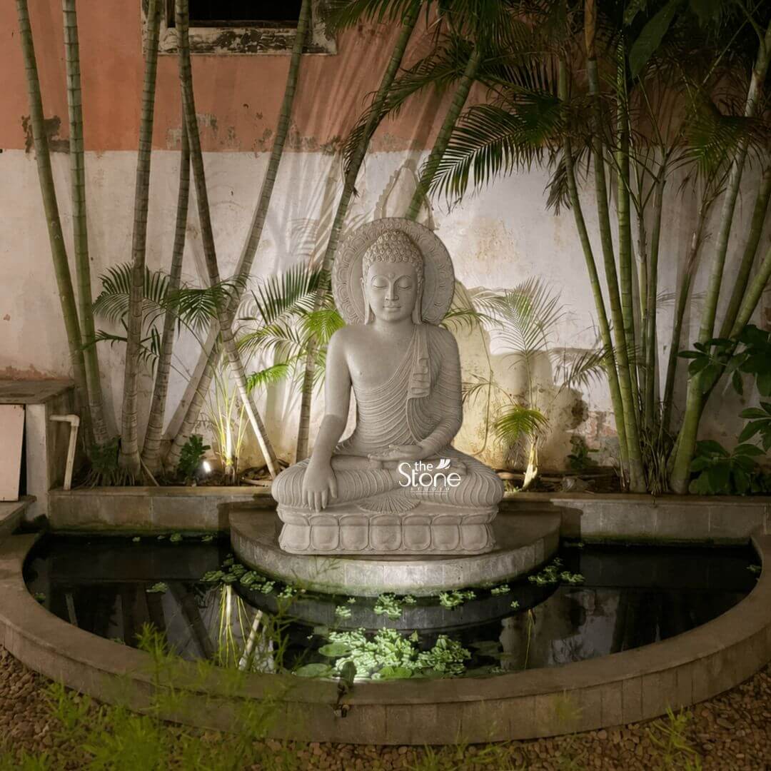 4 feet Buddha Statue 3 A magnificent four-foot-tall Big Buddha Idol sits as the tranquil centerpiece of a secluded garden courtyard installed on a raised platform within a crescent-shaped reflecting pond. The statue meticulously hand-carved from beautiful off-white sandstone depicts the Buddha in a traditional meditative pose seated on a carved lotus throne with his left hand resting in his lap and his right hand extended in the bhumisparsha mudra fingers pointing down to touch the earth. His face exhibits a serene contemplative expression with downcast eyes and a gentle smile framed by tightly curled hair and a cranial ushnisha all rendered with the soft matte texture and light beige-to-grey tones characteristic of the sandstone material. The setting is an artfully composed outdoor space designed for reflection. The statue is positioned against a weathered peach-colored stucco wall that is dramatically illuminated by uplighting casting soft shadows and highlighting the statue's form. Behind the Buddha a dense grove of tal slender bamboo stalks and lush green palm fronds creates a natural secluded backdrop with several plants flanking the platform and bathed in the same warm light. The reflecting pond in the foreground contains lily pads and other aquatic vegetation mirroring the image of the Buddha and the surrounding foliage adding a layer of depth and tranquility to the scene. The ground surrounding the pond is covered in small loose pebbles completing the garden aesthetic. This photograph captures the powerful presence of a 4-foot Buddha statue in off-white sandstone making it an ideal illustration for an article discussing Big Buddha Idol for Garden Decor. The statue's substantial size ensures it functions as a commanding focal point while the natural stone texture harmonizes beautifully with the surrounding plant life. The thoughtful use of lighting and the incorporation of the reflecting pond create a sacred and peaceful atmosphere perfectly demonstrating how such a large-scale idol can transform a garden into a sanctuary for meditation and aesthetic appreciation. The detailed carving and harmonious integration of elements highlight its value as a piece of sacred art in a modern landscape.