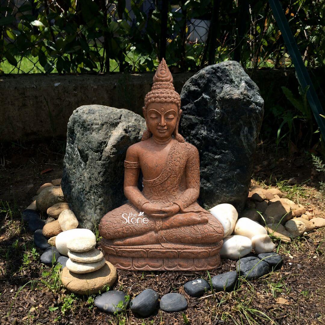 3.5 feet Garden Buddha Sculpture 3 A medium shot captures a majestic three-and-a-half-foot-tall Buddha statue hand-carved from a warm reddish-brown sandstone positioned as the spiritual focal point within an outdoor Zen garden. The statue is depicted in a traditional seated meditative posture known as Dhyana Mudra with both hands resting gently in the lap palms facing upwards and thumbs touching to form the mystic triangle. The Buddha's serene face conveys a profound sense of inner peace and stillness with eyes half-closed and a gentle smile. Its head is adorned with the characteristic snail-shell curls and the ushnisha protrusion representing wisdom and enlightenment. The sandstone sculpture exhibits a rich natural texture and detailed relief carving on the robe or sanghati which drapes elegantly over the left shoulder. The Buddha statue rests on a low raised stone plinth providing a stable and sacred foundation. The surrounding garden area is meticulously arranged with natural elements to create a tranquil sanctuary. Immediately behind the deity two large dark rugged natural stones serve as a striking backdrop providing a sense of grounding and stability against the softer hue of the sandstone. The statue's base is encircled by a careful arrangement of smooth river pebbles in contrasting colors a small stack of flat light-colored stones and a cluster of larger smooth white stones to the right and a scattering of larger dark grey pebbles in the foreground defining the sacred space. The setting is a lush sunlit corner of an outdoor garden with a natural earth and grass ground cover that includes small weeds and a few patches of mulch. Behind the arrangement of stones a dense dark green privet hedge creates a natural screen, isolating the Zen space from the rest of the garden. Natural daylight filters through the leaves above casting soft dappled shadows that emphasize the contours of the statue and the rich textures of the natural stone elements. The composition is balanced and inviting perfectly illustrating how a Dhyana Buddha statue can enhance the spiritual atmosphere and aesthetic of a Zen garden.