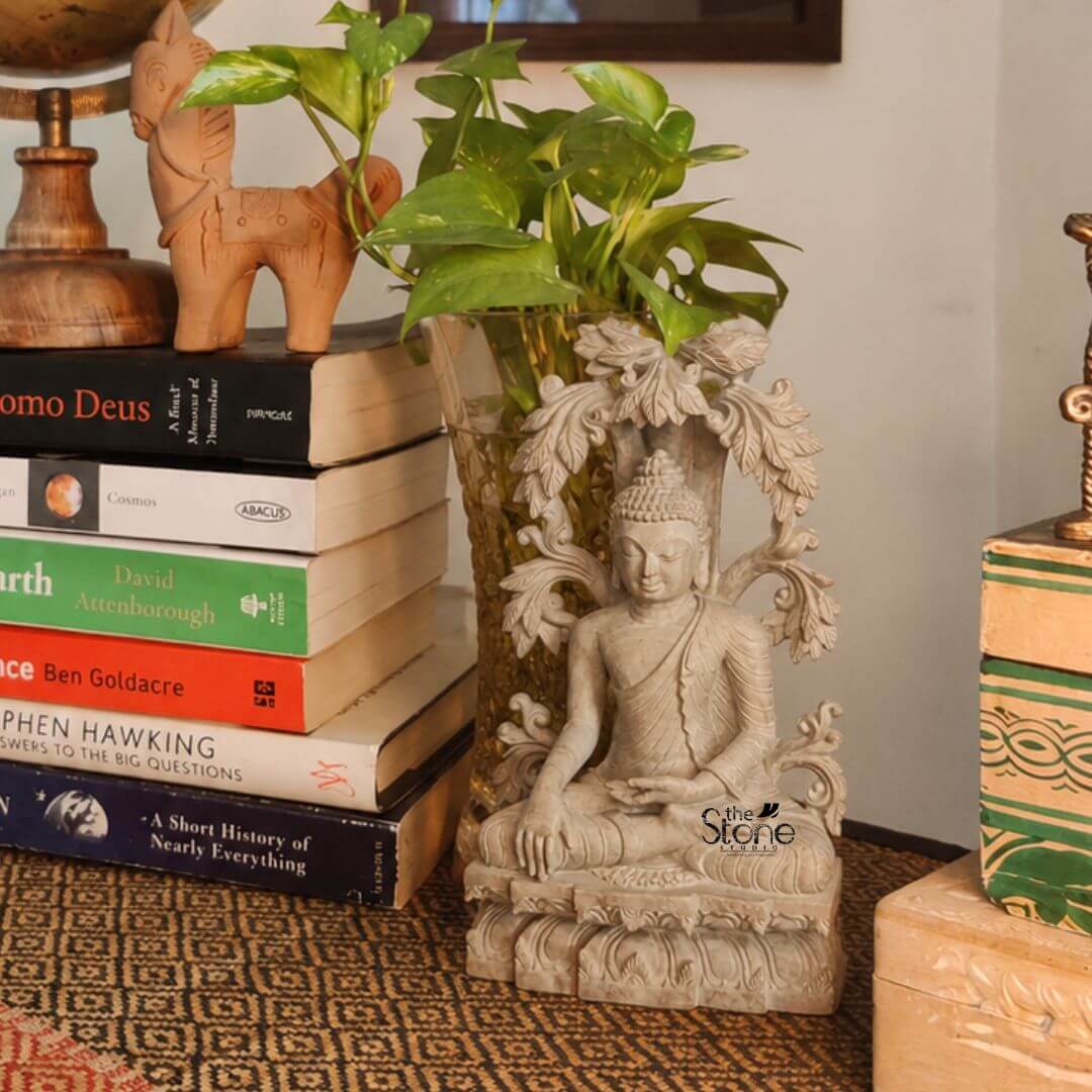 Small Buddha Statue For Home 7" 3 A small 7-inch decorative Buddha soap stone statue is styled as an elegant tabletop décor piece in a warm indoor setting. The handcrafted Buddha is seated in a calm meditative posture on a carved base with one hand resting on the lap and the other lowered in a traditional gesture. The figure features a serene expression closed eyes detailed robe folds curled hair and elongated earlobes while an ornate arch of flowing leaf and vine motifs rises around the statue adding artistic depth and a graceful sculptural frame. The Buddha statue is placed on a patterned table surface beside a neat stack of books helping show its compact size and suitability for table top décor. Behind the statue a clear glass vase filled with green money plant leaves adds freshness and a soothing natural touch. On the left wooden decorative objects and a small horse figurine rest above the stacked books while on the right a few more books and a neutral-toned decorative box complete the cozy cultured arrangement. The soft lighting and earthy colour palette give the scene a peaceful thoughtful, and refined atmosphere. This 7-inch soap stone Buddha statue is presented as an ideal small decorative Buddha for table top décor perfect for bookshelves study tables office desks, side tables meditation corners and living room styling. The image highlights how a compact handcrafted Buddha sculpture can blend spirituality with everyday interior design bringing serenity balance and timeless elegance to small spaces. Its detailed carving natural stone finish and tasteful placement make it a beautiful choice for home décor mindful interiors and subtle spiritual decoration.