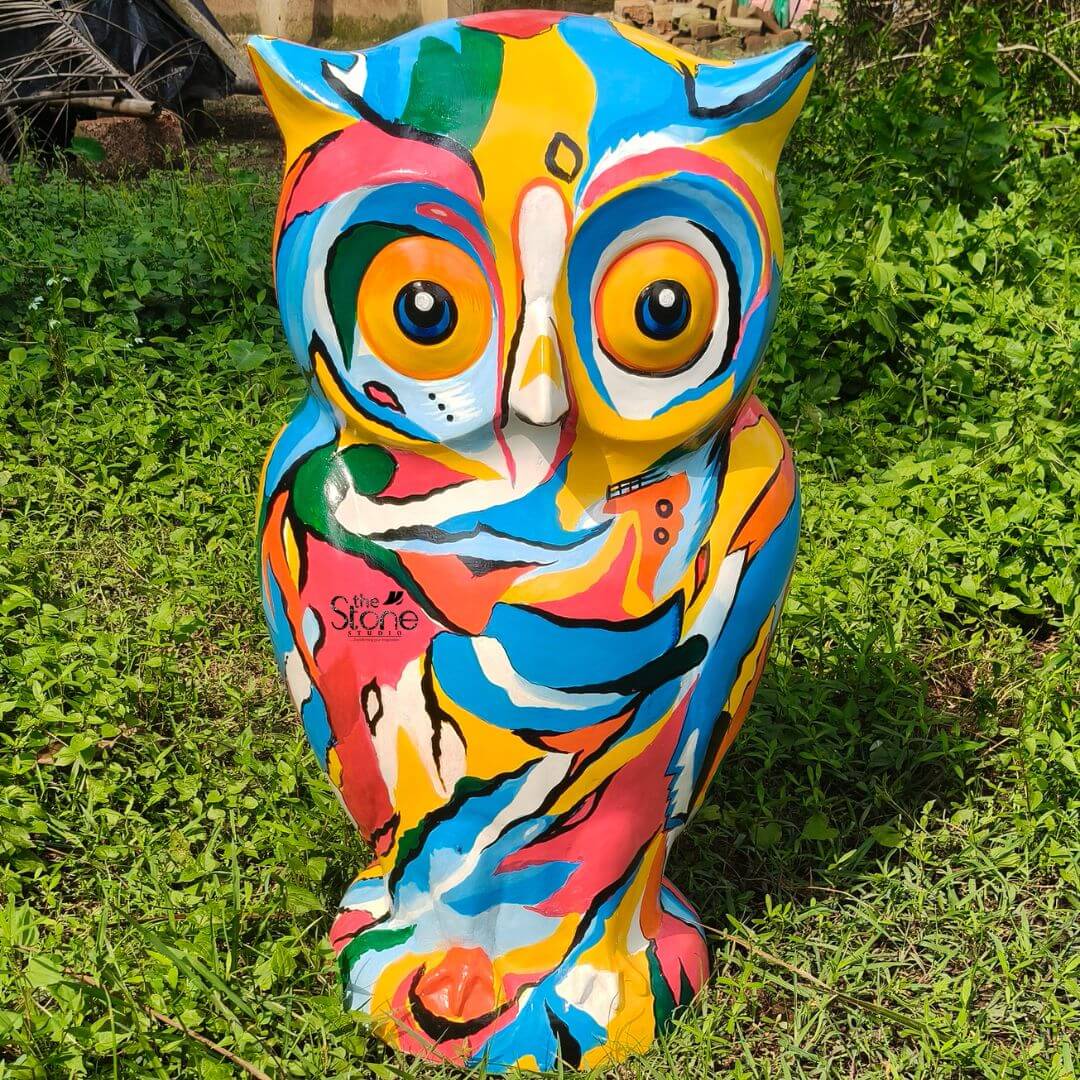 Pop Art Owl Statue 3ft 3 A vibrant three-foot-tall Fibre Pop Art Owl statue stands proudly outdoors nestled within a lush green patch of garden grass and weeds under bright sunlight. The sculpture is a kaleidoscope of color hand-painted with bold abstract swirling patches of hot pink turquoise blue sunny yellow orange and green. This dynamic non-representational pattern covers the entire surface transforming the realistic form of the owl into a striking piece of contemporary outdoor art. The statue is sculpted in a classic standing owl pose with prominent ear tufts and large wide-set eyes that are intricately incorporated into the colorful abstract design. The large engaging eyes feature orange irises and dark pupils outlined in white and yellow giving the owl a whimsical and captivating gaze. The surface of the statue has a smooth painted finish that catches the sunlight highlighting the rich layers of paint. The abstract color blocks flow into one another without defined outlines creating a fluid energetic pattern that wraps around the owl's head, chest and wings suggesting plumage without being strictly illustrative. The sturdy fibre material gives the statue a solid durable presence making it well-suited for a permanent outdoor display. This 36-inch Colorful Owl Garden Statue is designed to be a commanding and cheerful focal point in any landscape. Its large scale ensures it stands out against natural foliage while the Pop Art aesthetic brings a modern artistic flair to the garden. The statue's placement on the grass surrounded by the texture of plants perfectly illustrates how it functions as an outdoor owl garden sculpture adding personality and a burst of color to the natural environment. It is a perfect decorative piece for garden enthusiasts looking to add unique and eye-catching art to their outdoor living spaces.
