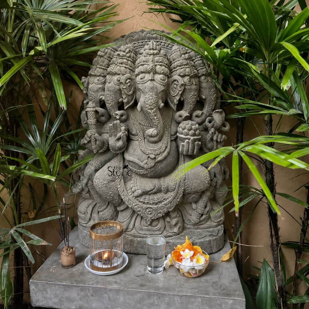 This is a focused view of a richly detailed, 32-inch-tall Panchamukhi Ganesha statue hand-carved from a dark textured blackstone. The statue is the central deity in a serene home altar setting, nestled among lush green tropical foliage that frames the sculpture against a warm textured wall. Ganesha is depicted in a seated, meditative posture featuring five distinct elephant heads facing in different directions with the central head looking forward and adorned with an elaborate crown. His ten arms hold various symbolic attributes including an axe, a goad, a lotus, and a moda all carved with intricate precision from the stone. The Panchamukhi Ganapati statue is seated upon a decorative stone plinth which rests on a modern smooth grey concrete pedestal. This setup creates a dedicated sacred space perfect for home decor or a meditation area. The dark matte finish of the blackstone provides a strong contrast against the vibrant greenery and the neutral wall emphasizing the fine craftsmanship and the complex iconography of the five faces. The statue is deeply carved with traditional jewelry, garlands and a sacred thread, showcasing the artisan's skill in rendering elaborate details in the hard stone. In the foreground on the concrete pedestal, are several items of offering arranged neatly a small lit oil lamp within a brass and glass cage a small glass of water and a decorative bowl filled with fresh yellow and white marigold flowers. These offerings enhance the devotional atmosphere and suggest the statue's active role within the home environment. Natural daylight filters in from the left casting gentle shadows that highlight the three-dimensional quality of the carving and the varied textures of the stone, plants and offerings. The overall composition presents a harmonious and spiritual focal point suitable for indoor worship or decoration.