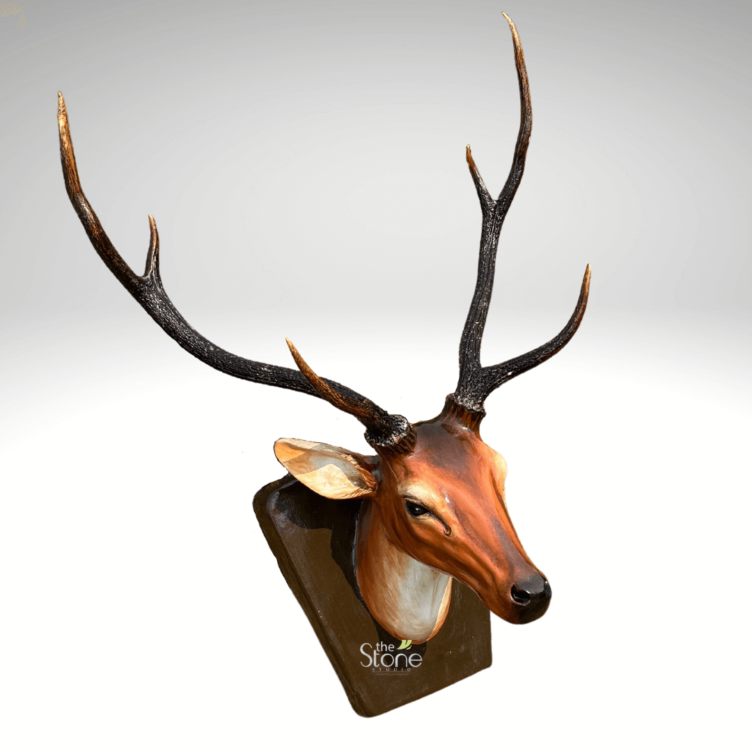 Deer Head Statue 2ft 3 highly detailed and lifelike three-quarter view of a faux deer head sculpture, meticulously crafted and mounted on a dark, polished plaque, presented against a clean, bright gradient background transitioning from light grey to white. The stag's head is rendered in rich, warm tones of reddish-brown and russet, smoothly transitioning to lighter cream and tan hues on its throat and underbelly, mimicking natural fur patterns. The deer's intelligent, dark eyes are particularly expressive, and a subtle, tear-like streak or artistic detail is visible just below the outer corner of its left eye, adding a poignant touch. Its ears are alert and slightly cupped, revealing lighter tan interiors. Dominating the top of the sculpture are the impressive, dark brown to black antlers, which feature a realistic, gnarled texture at their base where they emerge from the head. Each antler gracefully branches into multiple points or tines, with the tips featuring a lighter, almost golden-brown sheen. The entire head is secured to a sturdy, rectangular plaque with a dark brown, wood-like finish, angled slightly for optimal display. At the bottom center of the plaque, the white logo "The Stone Studio" is clearly visible, identifying the creator of this exquisite piece. This elegant wall-mounted deer head art offers a sophisticated and humane alternative to traditional taxidermy, ideal for enhancing rustic, lodge-style, or nature-inspired home decor and interior design themes.