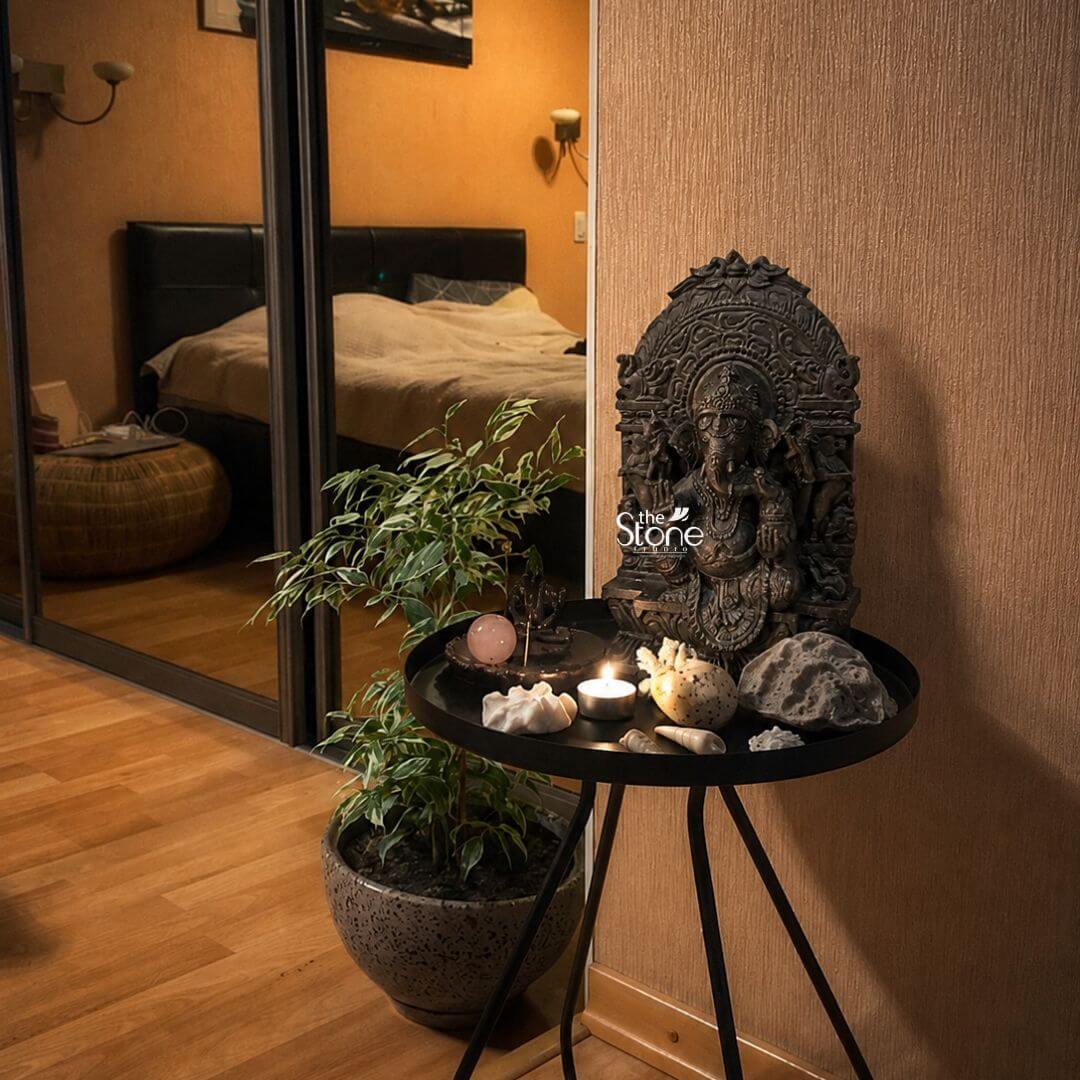A detailed close-up photograph capturing a beautifully styled home altar corner dedicated to Lord Ganesha. The central focus is a magnificent 15-inch tall Ganesha statue hand-carved from deep polished black stone. The statue depicts the elephant-headed deity in a seated posture upon a traditional pedestal with intricate carvings of an ornate archway rising behind him. His multiple arms hold symbolic attributes and he is adorned with detailed traditional jewelry and a crown. The dark smooth surface of the black stone reflects the ambient light highlighting the exceptional craftsmanship and fine details of the carving showcasing its spiritual significance. The Ganesha statue rests on a round black metal side table with a minimalist three-leg design placed against a textured light beige wall. The table is thoughtfully arranged as an offering space featuring various spiritual items that complement the idol. These include a small lit tea light candle casting a warm glow a polished rose quartz sphere a small crystal cluster various smooth sea shells and a larger porous gray volcanic rock formation. To the left of the table stands a potted Ficus benjamina plant with vibrant green leaves in a decorative gray speckled planter adding a touch of nature and freshness to the sacred space. The background of the image includes a large mirror with a dark frame which reflects the adjacent bedroom including a made bed with a beige duvet and a wicker pouf. This reflection adds depth and context showing the statue as an integrated part of a lived-in home. The warm directional light from the wall-mounted lamps in the bedroom illuminates the Ganesha statue and the surrounding objects creating a peaceful and inviting ambiance. The overall composition emphasizes the beauty and reverence of this small black stone Ganesha statue as a perfect centerpiece for any home temple or sacred corner.