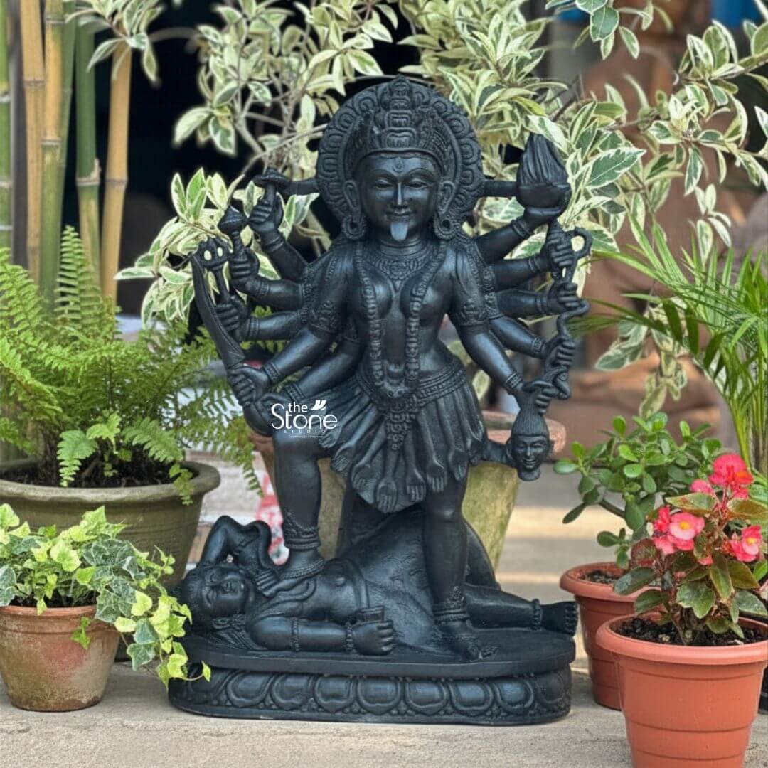 Kali Maa Murti Online 2ft 3 A powerful 2 feet Mahakali Murti in Black Stone meticulously carved from dark blackstone stands dramatically amidst lush greenery highlighting its fierce yet protective form. This 2 feet blackstone Maa Kali statue depicts the formidable goddess Mahakali embodying her destructive and transformative power. She is shown in her iconic stance with her right foot placed upon the reclining figure of Lord Shiva a symbolic representation of divine energy and consciousness. The smooth polished blackstone gives the statue a commanding and ancient presence beautifully contrasting with the vibrant plants. Maa Kali is depicted with multiple arms each holding a different weapon or symbolic item signifying her various powers to destroy evil and ignorance. Her tongue protrudes menacingly and her face though fierce also conveys a sense of divine purpose. She wears a crown necklaces and a short skirt all intricately carved in the blackstone consistent with traditional iconography. A halo or aureole behind her head further emphasizes her divine status. The intricate details of her jewelry weapons and her primary face showcase exceptional craftsmanship making it a compelling Mahakali Murti in Black Stone. The reclining figure of Lord Shiva beneath her foot is also carved with clear detail serene even in this powerful pose representing the balance of cosmic forces. The entire sculpture rests on a multi-tiered lotus-petal shaped base, also made of blackstone providing a sturdy and dignified foundation. The statue is surrounded by various potted plants including ferns iv and flowering plants with pink blooms creating a verdant and dynamic outdoor setting. In the background taller plants like bamboo add depth. The overall composition powerfully conveys Mahakali's immense strength her role as a destroyer of evil and her ultimate protective nature. This detailed description effectively connects the visual elements of the blackstone sculpture with its spiritual significance and aesthetic qualities.
