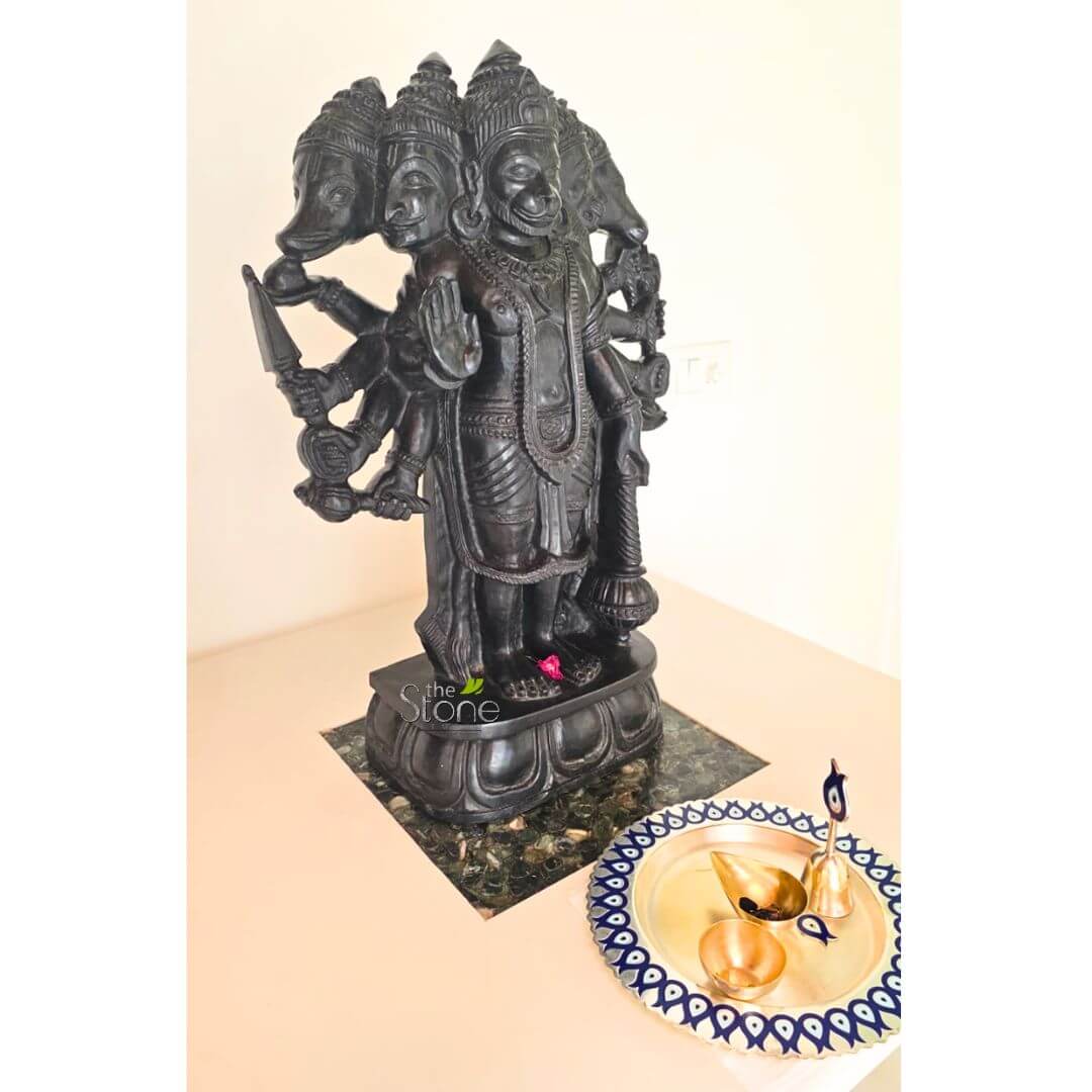 Panchmukhi Hanuman Stone Statue 2ft 3 striking and tall black stone sculpture of Panchamukhi Hanuman, the five-faced form of the revered Hindu deity Hanuman, stands prominently as the central focus of the image. The idol is meticulously carved from a polished, dark stone, possibly granite or soapstone, showcasing intricate details in its craftsmanship. Hanuman's central face, depicting a benevolent and smiling expression, is adorned with an elaborate crown. It is flanked by four other distinct heads, representing specific divine forms: on the far left (from the viewer's perspective) is the head of a boar (Varaha), and on the immediate left, a lion's head (Narasimha). The other two heads, completing the traditional Panchamukhi iconography, appear to be a horse (Hayagriva) and a mythical bird (Garuda), though their finer details are partially obscured by the statue's dark material. The deity is depicted with multiple arms, at least eight clearly visible, each holding various divine attributes or weapons. On the left side, Hanuman grasps a sharp dagger or sword, a bow, and a heavy mace (gada). On the right side, he holds another mace and extends an open palm in the Abhaya Mudra, a gesture signifying fearlessness and protection. The sculpture is further adorned with finely carved necklaces, armbands, and a long garland draped around its torso. A solitary, vibrant pink flower rests delicately at the base of the deity's feet. The sculpture stands on a multi-tiered lotus pedestal, which itself rests on a dark, mottled green and black stone slab. This entire devotional display is set upon a smooth, light beige or cream-colored tabletop. To the right of the statue, a beautiful golden brass pooja thali (ritual plate) is positioned. Its rim features an elegant, repeating pattern of dark blue, eye-shaped or stylized wave motifs. Inside the thali are several items typically used in Hindu worship: a small, ornate brass bell with a dark blue evil eye charm attached to its handle, a small, boat-shaped brass oil lamp (diya), and a tiny empty brass bowl. The background is a plain, light-colored or white wall, creating a clean and reverent setting that highlights the spiritual significance of the artifacts. This detailed depiction suggests a cherished home altar, a display of religious art, or an artifact for sale, emphasizing traditional Indian craftsmanship, spiritual devotion, and the rich iconography of Hindu mythology.