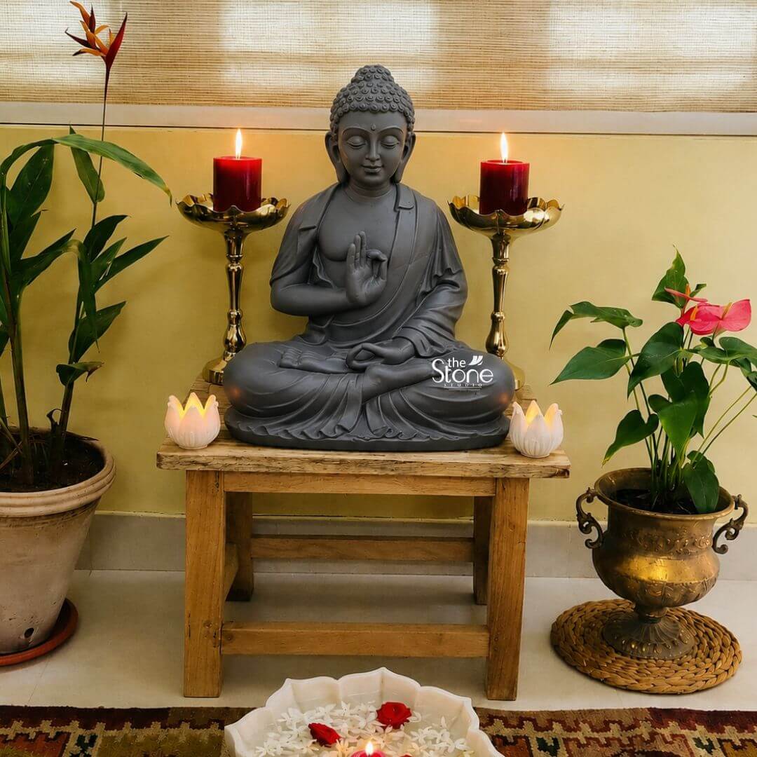 A full-view photograph captures a serene indoor home altar setup centered around a magnificent 24-inch tall Fibre Buddha Idol. The Buddha statue rendered in a dark matte gray finish that mimics stone is seated in the lotus position upon a rustic wooden stool embodying profound tranquility. His hands are held in the vitarka mudra symbolizing the transmission of teaching and discussion with the thumb and index finger touching to form a circle. The idol exhibits a calm facial expression with downcast eyes and the traditional curled hair and ushnisha showcasing the fine detail achievable with high-quality fibre construction. The statue is placed on a smal weathered wooden bench which serves as a makeshift altar against a plain warm cream-colored wall. Symmetrical elements frame the central figure, enhancing the sacred atmosphere. Two tall, ornate brass candlesticks, each holding a glowing red pillar candle, stand behind the Buddha. In front of the statue, two small, white lotus-shaped ceramic tea light holders with lit candles rest on the bench. To the left of the altar sits a large potted plant with broad green leaves in a simple beige planter, and to the right is a smaller, ornate antique brass urn-style planter containing a vibrant green plant with pink anthurium flowers, resting on a woven jute mat. In the immediate foreground, a shallow, square white ceramic bowl is filled with water, floating white jasmine flowers and red rose petals, with a single lit candle at its center. The entire composition creates a peaceful and inviting sanctuary for meditation and contemplation within a home setting. The placement of the idol, combined with the soft glow of the candles and the freshness of the plants, highlights its role as a beautiful and spiritual focal point for home decor. The use of fibre makes this detailed and substantial statue a durable and manageable piece for interior styling.