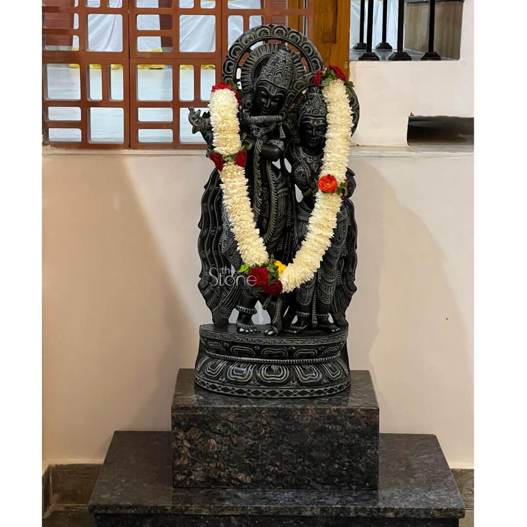 Radha Krishna Statue For Home 30" 3 majestic and intricately carved black stone statue of the divine Hindu couple, Lord Krishna and Radha, stands as the central focus of the image. The figures are depicted standing gracefully side-by-side on a decorative pedestal. Lord Krishna, positioned on the left, is shown with a serene expression, holding his signature flute to his lips, suggesting a moment of melodic devotion. Radha, on the right, stands lovingly beside him, gazing towards Krishna, with her arm gently around his waist or linked with his arm. Both deities wear elaborate crowns adorned with intricate details, traditional flowing drapery that reveals the contours of their forms, and delicate jewelry including necklaces, armlets, and waistbands, all meticulously sculpted from the dark, polished stone. Behind their heads, a stylized, circular arch or halo, also carved from the same material, adds to their divine aura. The craftsmanship highlights fine details in their facial features, garments, and accessories, showcasing skilled artistry. Adorning the statue are two thick, fresh garlands of white, fragrant jasmine-like flowers, generously interspersed with vibrant red roses and yellow blossoms. These garlands are draped in a classic 'V' shape from the deities' shoulders, extending down to their mid-sections, symbolizing reverence and offering. The Radha Krishna idol rests on a multi-tiered pedestal. The statue's immediate base is a rounded, intricately carved dark stone lotus-petal design, which sits atop a larger, rectangular block of dark, speckled granite with subtle grey and brown variations. This main pedestal is further supported by an even wider, lower matching granite base, providing a substantial and elegant foundation. The background features a light, off-white wall, suggesting an indoor setting like a home puja room, temple niche, or a gallery showroom. In the upper left quadrant, a distinctive brown wooden lattice screen or window with a geometric pattern is visible, allowing a soft, diffused light into the space. The overall composition conveys a sense of spiritual tranquility, cultural heritage, and exceptional traditional Indian stone carving, perfect for discussions about devotional art, spiritual home decor, or the craftsmanship of Hindu deity sculptures. The presence of the "5th Stone" watermark subtly suggests the origin or craftsmanship of this beautiful black granite Krishna Radha statue.