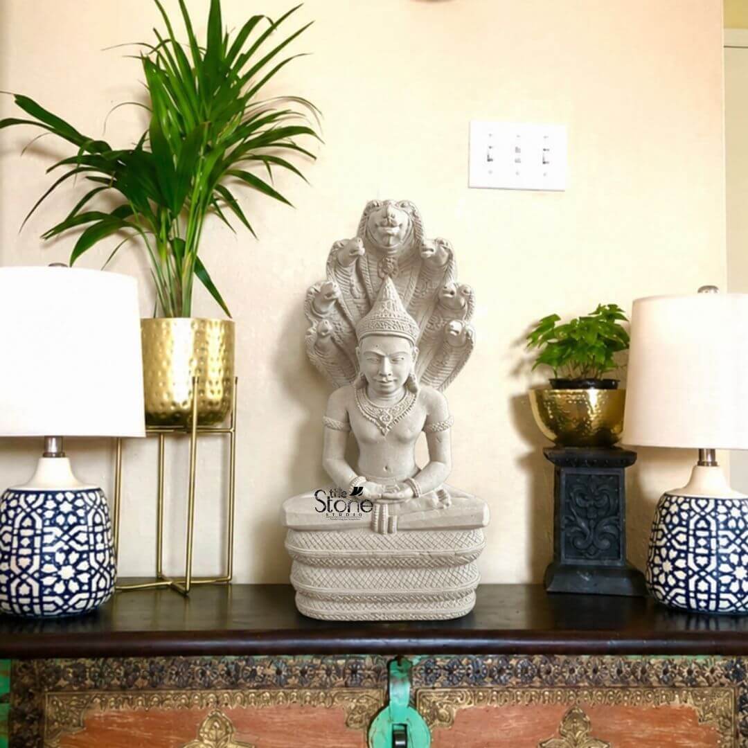 Angkor Wat Style Meditation Buddha 18" 3 An 18-inch white sandstone Buddha idol is displayed as the centrepiece of a well-styled home décor arrangement placed on a dark wooden console against a soft cream wall. The sculpture shows Buddha seated in a calm meditative posture on a detailed coiled pedestal with hands resting gently in the lap and a serene facial expression that conveys peace and stillness. Behind the figure rises a dramatic multi-headed serpent canopy creating a striking and sacred silhouette that gives the statue a unique and artistic presence. The fine carving on the crown jewellery facial features and pedestal adds richness to the white sandstone finish making the idol look both devotional and decorative. The Buddha statue is framed by a balanced interior setting that enhances its appeal as a unique Buddha idol for home décor. On both sides of the sculpture are table lamps with white shades and blue patterned ceramic bases creating visual symmetry and a warm homely atmosphere. To the left a tall green indoor plant in a hammered gold planter adds height and freshness while on the right a smaller leafy plant in a matching metallic planter sits elevated on a dark carved stand. The contrast of greenery gold-toned planters patterned lamps and the pale sandstone statue brings texture warmth and elegance to the overall arrangement. This image clearly connects to an article about a Unique Buddha Idol for Home Decor by showing how an 18-inch white sandstone Buddha statue can serve as a meaningful focal point in a living room entrance area meditation corner or console table display. The combination of the meditative seated Buddha the ornamental serpent backdrop and the refined indoor styling highlights both spiritual symbolism and decorative charm. The statue appears ideal for those seeking a handcrafted sandstone Buddha idol that adds serenity character and timeless beauty to home interiors while also standing out as a distinctive and elegant décor piece.
