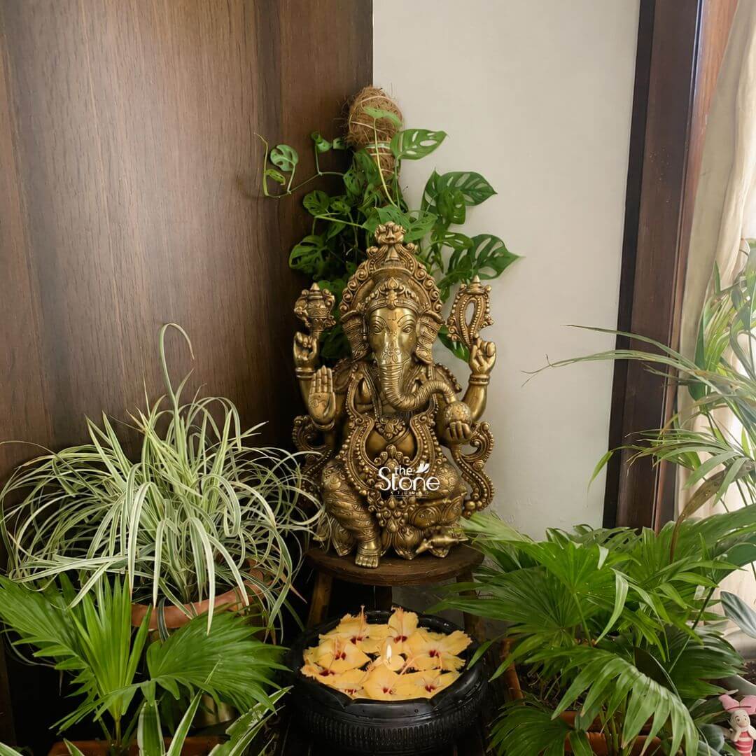 Brass Ganesh Murti 21" 3 A 21 inch brass Ganesha idol is displayed as the central feature of a calm and beautifully styled home décor corner. The statue depicts Lord Ganesha seated in a traditional posture with four arms a curved trunk large ears and a serene welcoming expression. The brass finish has a warm antique golden tone that enhances the richness of the sculpture while the detailed crown jewellery sacred ornaments and decorative throne-like backrest highlight the fine craftsmanship. One hand is raised in a blessing gesture while the other hands hold symbolic objects adding devotional significance to the elegant brass Ganesha statue. The Brass Ganesha idol is placed on a small dark wooden stool and surrounded by lush green indoor plants creating a refreshing and harmonious setting. Behind the statue climbing and leafy plants rise against a wooden wall panel while potted spider plants and palm-like foliage fill the foreground and sides. At the base of the arrangement is a dark bowl filled with water and floating yellow flowers adding a soft spiritual and decorative touch. Natural light entering from the side brightens the plants and the brass surface making the home décor setup feel peaceful warm and inviting. This image connects perfectly to an article about a Brass Ganesha Idol For Home Decor Statue by showing how a 21 inch brass Ganesha statue can become a graceful spiritual focal point in a living room corner entrance area balcony nook meditation space or home temple setting. The combination of antique brass greenery natural textures and floral accents demonstrates how a Ganesha idol for home décor can blend devotion with elegant interior styling. The overall arrangement conveys blessings prosperity positivity and timeless decorative appeal making this brass Ganesha statue a meaningful and stylish choice for home décor.