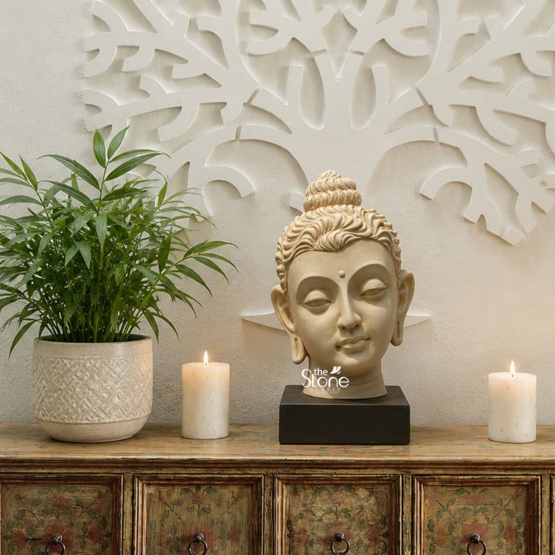 Gandhara Art Buddha 1ft 3 A serene 12-inch Buddha Head Statue meticulously carved from soapstone is elegantly positioned on a dark rectangular base serving as a tranquil centerpiece for tabletop decor. The Buddha head with its finely sculpted features embodies a sense of deep inner peace and spiritual wisdom. His eyes are gently closed in meditation with a subtle smile on his lips and elongated earlobes indicative of his princely past. The light creamy hue of the soapstone gives the Buddha head a soft and inviting presence blending harmoniously with the sophisticated decor. The hair is intricately sculpted in tight curls culminating in the ushnisha the cranial bump symbolizing enlightenment. The Buddha head is placed on a beautifully distressed antique-style wooden console table with painted floral or abstract patterns and ornate metal handles. Flanking the statue are two unlit pillar candles adding to the calm and contemplative ambiance. This 12-inch soapstone Buddha Head statue is an ideal subject for an article on Buddha head statues for tabletop decor. To the left of the statue a lush green potted plant in a decorative white ceramic pot adds a touch of natural freshness. In the background a striking white carved or relief wall art piece possibly depicting a tree or abstract organic shapes complements the spiritual theme. The entire arrangement creates a harmonious and aesthetically pleasing display perfect for bringing a sense of calm mindfulness and spiritual elegance to any home interior.