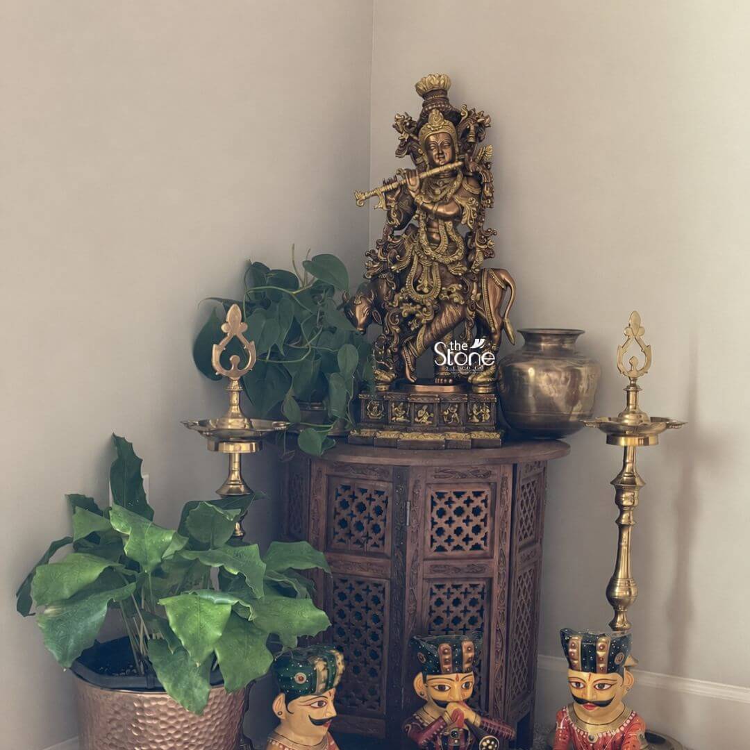 Krishna With Cow Brass Idol 28" 3 A 28 inch brass Krishna idol is displayed as the central feature of a beautifully arranged home décor corner creating a devotional and elegant focal point. The statue depicts Lord Krishna in a graceful standing posture holding a flute across the body in the classic divine musician form. The idol has a richly detailed crown expressive facial features layered ornaments and flowing decorative elements that give the sculpture a regal and sacred presence. The warm antique brass finish highlights the intricate craftsmanship and gives the Krishna statue a timeless traditional look well suited for spiritual home styling. The brass Krishna statue is placed on top of a carved wooden cabinet with lattice-style doors positioned neatly in the corner against soft neutral walls. On both sides of the idol stand tall traditional brass oil lamps adding symmetry and reinforcing the devotional atmosphere. A rounded metal vessel sits beside the statue while trailing green pothos leaves behind the idol add freshness and softness to the arrangement. In the foreground a large leafy plant in a textured metallic planter and a pair of colourful decorative figurines bring extra character warmth and visual richness to the overall interior setting. This image connects perfectly to an article about a Brass Krishna Idol for Home Decor by showing how a 28 inch brass Krishna statue can become a striking spiritual and decorative centrepiece in a living room prayer corner foyer or meditation space. The combination of the ornate brass idol wooden furniture traditional lamps indoor plants and handcrafted accents demonstrates how a Krishna idol for home can blend devotion with tasteful interior décor. The overall scene conveys serenity elegance cultural richness and the timeless beauty of a handcrafted brass Krishna statue designed to bring divine charm and artistic appeal into the home.