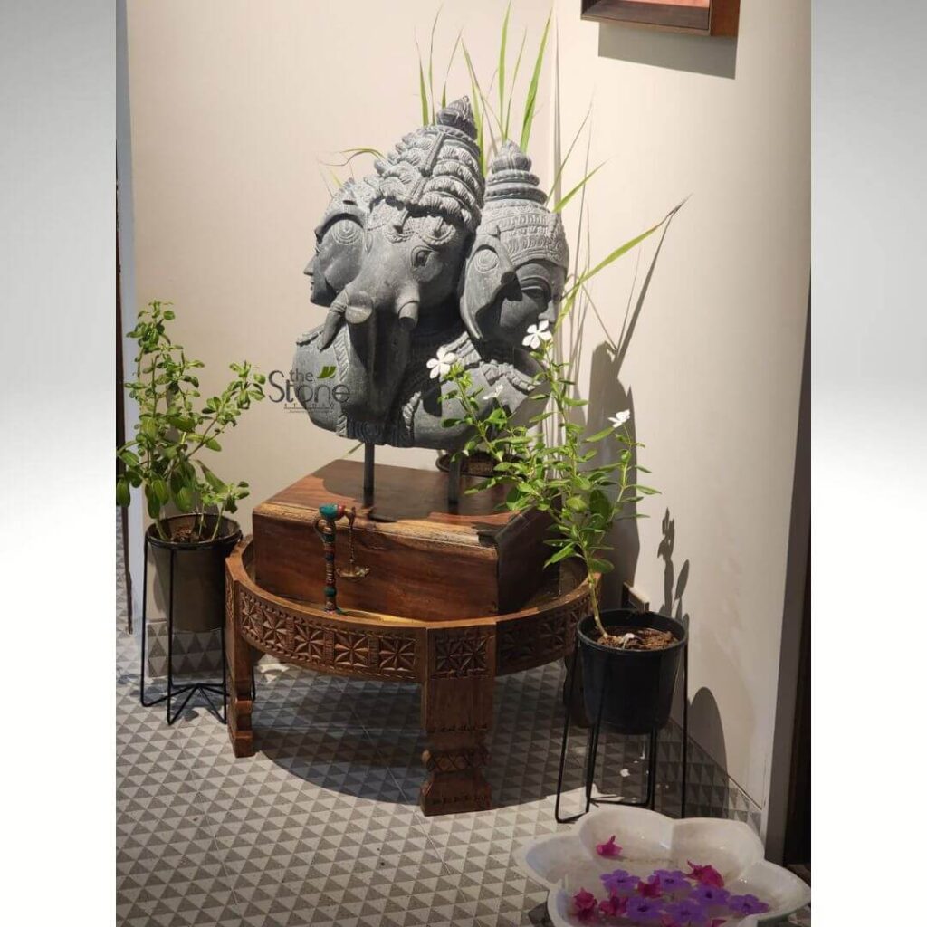 Decorating Apartment with small Statues 17 captivating indoor vignette showcases a large, intricately carved dark grey stone sculpture depicting three divine heads, placed as a focal point in a well-styled corner. The central and most prominent head is that of Lord Ganesha, easily identified by his elephant trunk, large ears, and elaborate crown, exuding a sense of wisdom and benevolence. He is flanked on either side by two human-like busts, likely deities or revered figures, each adorned with detailed traditional Indian headdresses and serene facial expressions. The entire multi-headed sculpture rests on a sleek black metal stand, which in turn is positioned atop a substantial, circular wooden table crafted from rich, dark polished wood. The table features exquisite hand-carved geometric patterns along its perimeter, highlighting traditional craftsmanship. Surrounding this spiritual centerpiece are several lush green potted plants, adding natural vibrancy to the display. Tall, slender blades of grass rise behind the sculpture, while smaller plants with delicate white blossoms are arranged in black and dark brown pots on elegant metal stands on either side. A small decorative charm, resembling miniature scales with colorful beads, hangs from the side of the wooden table. In the lower right foreground, a beautiful white, lotus-shaped ceramic bowl holds clear water with an assortment of vibrant purple and pink flower petals gently floating on its surface, creating a tranquil and inviting atmosphere. The setting is completed by a contemporary grey and white patterned tile floor and a neutral, light-colored wall in the background, making the dark stone and wood elements truly stand out. This image beautifully illustrates the integration of traditional Indian spiritual art and high-quality artisanal home decor, likely from a specialist in stone craftsmanship, creating a serene and culturally rich interior space. The "the Stone" watermark subtly visible on the sculpture reinforces its origin or featured product status within the context of an article on exquisite stone carvings or decor, showcasing the artistry and craftsmanship of their spiritual statues and home accents.