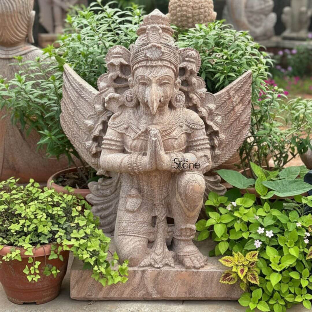 Stone Garuda Statue 2.5ft 6 A striking 2.5-foot sandstone Garuda statue perfect for home decor depicted kneeling in a posture of reverence amidst a vibrant garden setting as featured in an article about such sculptures. The entire statue is carved from natural reddish-brown sandstone giving it an earthy and textured appearance. Garuda the mythical bird-like vehicle of Lord Vishnu is depicted with a human-like body and a prominent beaked face. His hands are joined in Anjali Mudra symbolizing devotion and respect. The intricate details of the carving are evident in Garuda's elaborate crown the feathered wings spread elegantly behind him and the traditional jewelry adorning his neck chest, and arms. His powerful muscular legs are bent in a kneeling posture suggesting readiness for flight or deep contemplation. The texture of the sandstone is visible throughout adding to the rustic charm and authenticity of the sculpture. The sculptor has meticulously rendered the feathers of his wings and the folds of his lower garment showcasing exceptional artistry. The statue is placed on a simple rectangular sandstone base surrounded by various potted plants and lush green foliage creating a serene and natural ambiance. Hints of other stone sculptures are visible in the blurred background suggesting a larger collection or garden display. This 2.5-foot sandstone Garuda statue is a powerful representation of devotion strength and mythical grandeur making it an ideal piece for enhancing the spiritual and aesthetic appeal of any home or garden space as discussed in the accompanying article.
