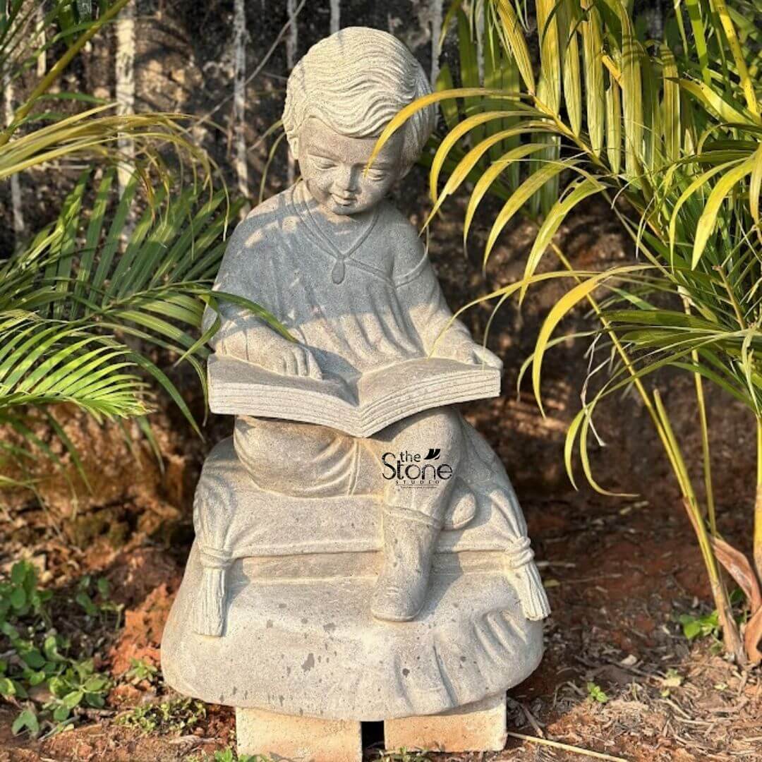 Reading Kid Statue 2ft 3 A delightful 2-foot Black Stone Reading Boy statue an ideal Reading Boy Stone Statue for Garden is charmingly situated amidst lush greenery in an outdoor setting. The statue carved from dark black stone depicts a young boy sitting cross-legged engrossed in reading an open book. His hair is neatly styled and he wears a simple tunic and a small necklace giving him an innocent and studious appearance. The detailing on the boy's face showing a focused gentle expression as he looks down at his book is quite engaging. The boy is seated on a plush-looking cushion or pillow sculpted with details like tassels at the corners adding a touch of comfort to his pose. The open book in his hands is also meticulously carved suggesting pages and a binding. The dark black stone of the statue provides a striking contrast against the vibrant green foliage surrounding it making the figure a prominent and endearing focal point in the garden. Despite being described as black stone in the prompt the image shows a greyish stone which still provides a beautiful contrast with the greenery. The statue is positioned on a small rustic pedestal partially hidden by the surrounding plants and earth. It is flanked by tall leafy plants including palm-like foliage creating a serene and natural backdrop. This 2-foot Black Stone Reading Boy statue is an exquisite example of garden decor designed to inspire a sense of calm contemplation and perhaps a love for learning in any outdoor space perfectly aligning with an article focusing on such charming and contemplative garden sculptures.
