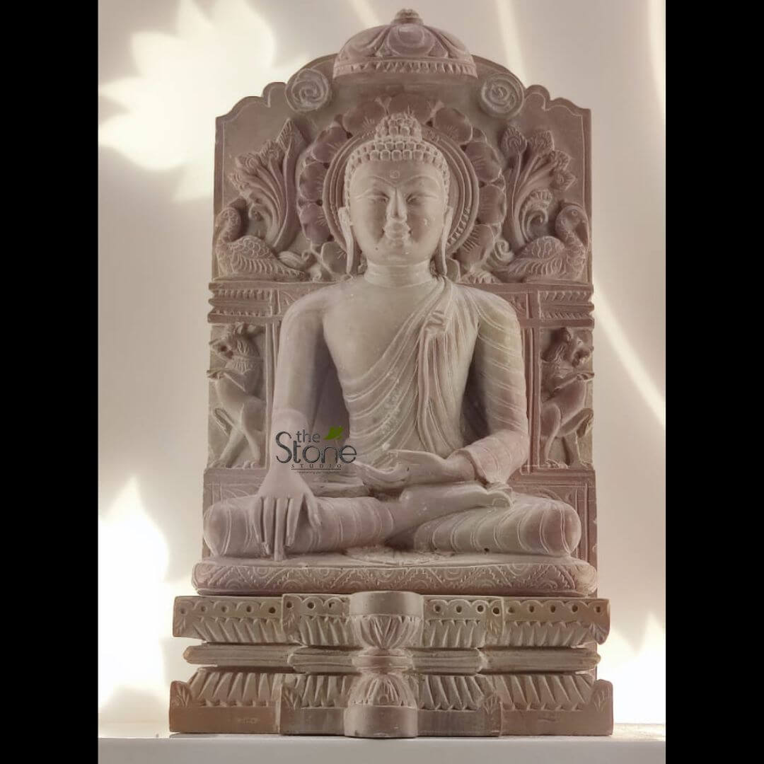 8 Inch Buddha Statue 3 meticulously hand-carved stone sculpture of Buddha, rendered in a warm, earthy-toned reddish-brown or pink sandstone, presented against a plain, brightly lit white background. The central figure of Siddhartha Gautama Buddha is depicted in a deeply meditative posture, seated in the full lotus position (Padmasana) with legs crossed, feet resting on the thighs, and soles visible. His hands rest gently in his lap, palms upwards, forming the Dhyana Mudra, symbolizing deep concentration, spiritual awareness, and the unity of mind and body. A serene and contemplative expression graces his face, characterized by downcast eyes, a gentle smile, a prominent urna (a dot between the eyebrows signifying spiritual insight), and elongated earlobes, indicative of his princely past before renunciation. His monastic robe is draped gracefully over his left shoulder, showing naturalistic, flowing folds and textures that highlight the skilled craftsmanship of the stone carving. Behind the Buddha, an intricately carved backslab, often referred to as a Prabhavali or Torana, frames the deity. It features a circular halo (prabhamandala) behind his head, adorned with a delicate, multi-layered lotus flower motif, symbolizing purity and spiritual growth. The sides of the backslab are richly ornamented with auspicious symbols and mythical creatures: on each side, a graceful swan or peacock-like bird with elaborate tail feathers is carved above a stylized deer or antelope, surrounded by vibrant scrollwork and lush foliage. The entire backslab is surmounted by an ornate, dome-like finial, adding to its architectural grandeur. The Buddha sits upon a multi-tiered pedestal, which is equally detailed with traditional architectural elements and decorative bands, including stylized lotus petals, geometric patterns, and intricate scrollwork, enhancing the overall beauty and cultural significance of the piece. A small, stylized support column is visible at the very front of the base. Soft, directional lighting illuminates the sculpture from the upper left, casting subtle shadows that highlight the textures, contours, and three-dimensional depth of the detailed carvings, imparting a profound sense of peace and tranquility. This traditional Buddhist artifact, showcasing exceptional stone craftsmanship and artistry, serves as a powerful symbol of meditation, spiritual practice, and inner peace, making it an ideal piece of spiritual art or timeless home decor. The watermark "the Stone Studio" is subtly visible on the statue's body, indicating the origin of this artisan piece.