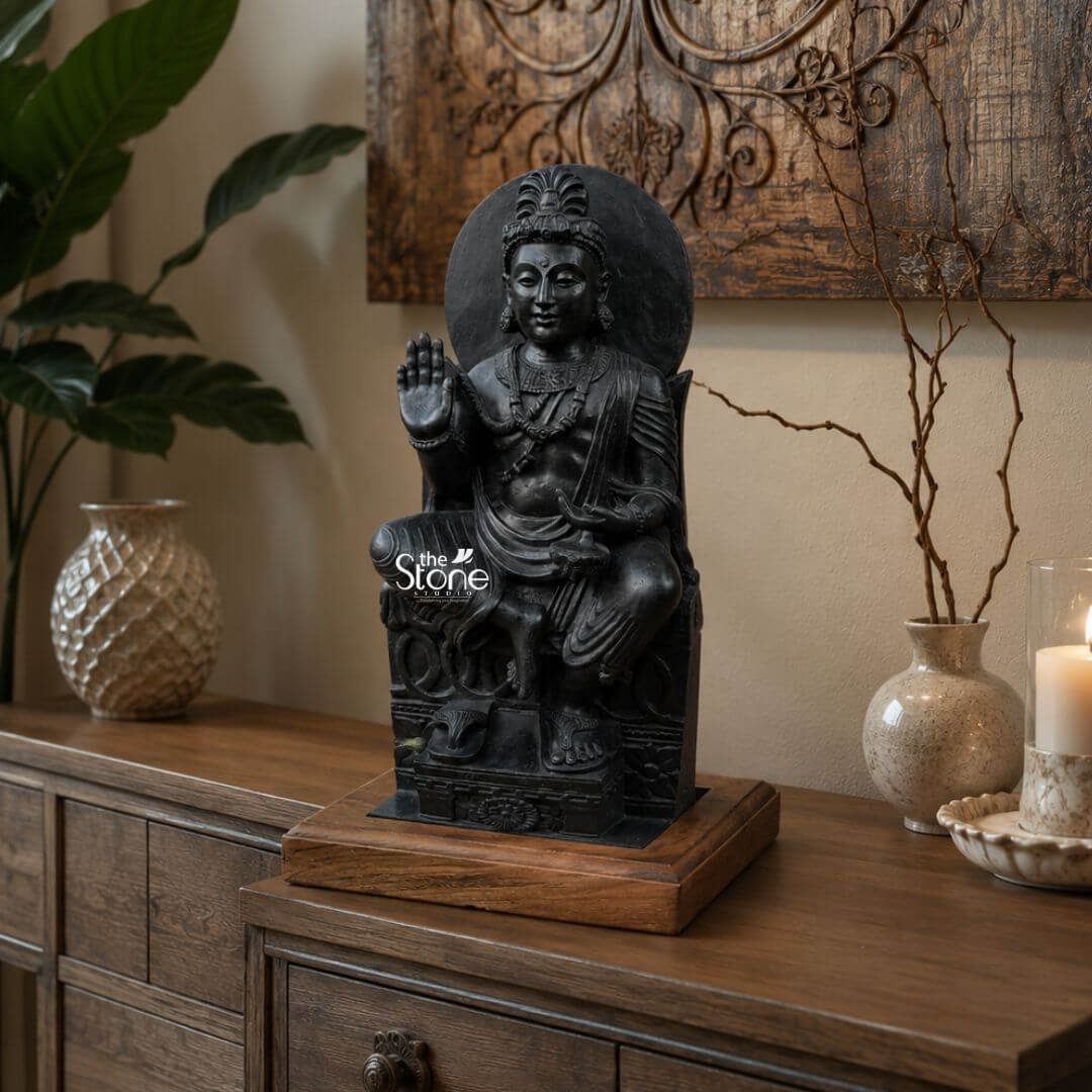 Avalokiteshvara Gandhara Statue 20" 3 A striking 20-inch Blackstone Avalokiteshvara Gandhara statue perfectly suited for table top decor commands attention on a rich wooden console table in a warmly lit interior. The statue is carved from dark black stone, giving it a profound and timeless appearance and depicts Avalokiteshvara the Bodhisattva of Compassion seated gracefully. His right hand is raised in a gesture of reassurance or blessing while his left hand rests gently on his lap, holding what appears to be a small offering or symbol. The Gandhara style is evident in the realistic drapery of his robes and the serene somewhat Hellenistic features of his face. Avalokiteshvara is adorned with an elaborate crown and intricate jewelry around his neck, chest and arms indicative of his divine status. A circular halo or aureole frames his head enhancing his spiritual presence. The statue is seated on an ornate throne-like structure with carved details and the overall craftsmanship showcases the intricate artistry possible with blackstone. The statue itself rests on a contrasting lighter wooden base which in turn sits on the polished surface of the console table elevating its display. The surrounding elements enhance the statue's appeal as a piece of table top decor. To the left a textured cream-colored vase and a large green plant add organic touches. To the right a pair of elegant cream-colored vases with subtle patterns one holding delicate dry branches flank a lit candle in a decorative holder casting a soft glow. A large intricately carved wooden wall art piece hangs above the table complementing the statue's cultural aesthetic. This 20-inch Blackstone Avalokiteshvara statue is a superb example of a Stone Buddhist Statue for Table Top Decor bringing a sense of ancient artistry spiritual calm and sophisticated elegance to any home.