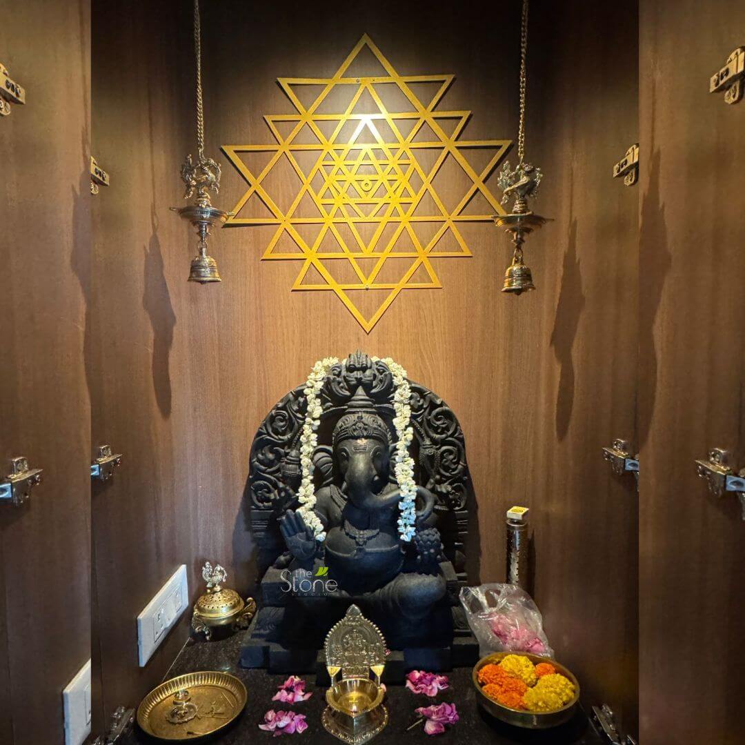 2 Feet Ganesh Statue 3 beautifully adorned and serene home pooja room or meditation space features a striking black stone idol of Lord Ganesha as its central focus. The deity is seated in a tranquil pose on an intricately carved matching black stone throne, with his trunk curving gently to the left. He is adorned with a fresh white floral garland, likely made of jasmine, draped elegantly around his neck and shoulders. One of Ganesha's hands is raised in the classic Abhaya Mudra, a gesture of blessing and fearlessness, while the other holds a traditional sweet, a modak or laddoo. The idol rests on a polished dark altar surface, which showcases various offerings and ritualistic items. To the left of Ganesha, a small, ornate brass plate and an intricately designed brass incense burner with a decorative finial are placed. Directly in front of the deity, an exquisite brass oil lamp (diya) sits amidst scattered pink and purple flower petals, suggesting recent or ongoing worship. To the right, a vibrant brass bowl overflows with fresh orange and yellow marigold flowers, symbolizing devotion and purity, alongside a clear plastic bag containing more pink blooms, ready for offering. The background consists of warm, dark brown wooden paneling, creating a cozy and intimate alcove. Dominating the wall directly above Ganesha is a magnificent golden Shri Yantra, a complex geometric diagram made of interconnected triangles, which subtly glows with a soft, ethereal light from behind, signifying divine energy and cosmic harmony. Flanking the Shri Yantra are two ornate brass hanging lamps (diyas), each suspended by delicate chains and featuring intricate detailing, including bell-shaped bottoms. These lamps cast elegant, elongated shadows on the wooden background, adding depth and visual interest to the sacred space. The overall composition evokes a strong sense of spirituality, tranquility, and devotion, characteristic of Hindu home shrines. This detailed image vividly portrays a dedicated space for worship and meditation, embodying tranquility and spiritual reverence, which could be central to an article discussing home spirituality, Hindu traditions, or interior design incorporating sacred spaces.