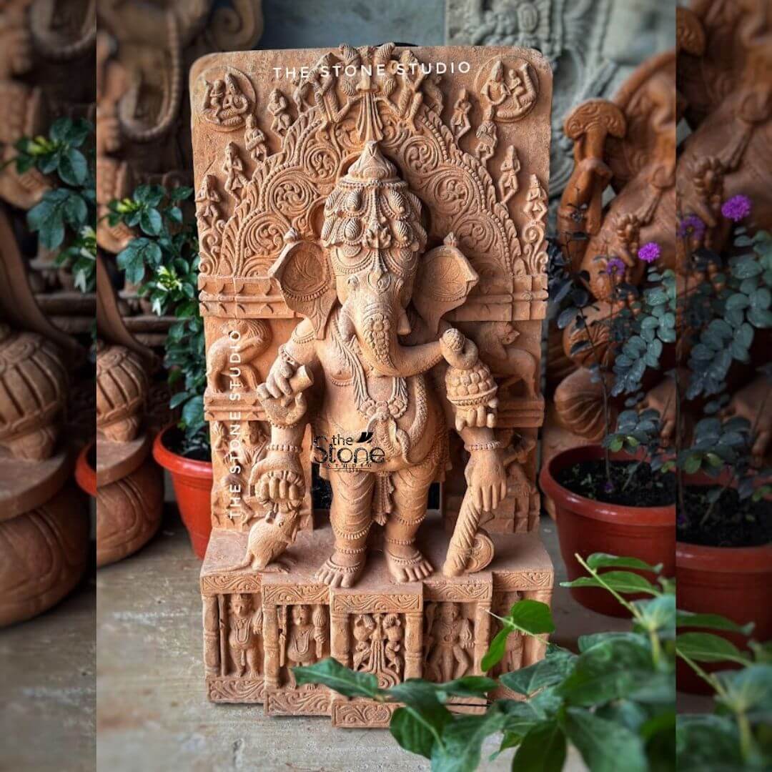 Ganesh Standing Statue 3ft 3 A magnificent 3-foot Brown Sandstone Ganesha statue perfectly suited as a Standing Ganesha Statue for Garden stands prominently in a vibrant outdoor setting. The statue carved from warm reddish-brown sandstone depicts Lord Ganesha in a dynamic standing posture radiating benevolence and divine energy. His iconic elephant head is intricately sculpted featuring large ears expressive eyes and his distinctive broken tusk. He is adorned with an elaborate crown traditional jewelry and flowing garments all carved with remarkable detail emphasizing the natural texture and beauty of the sandstone. Ganesha is portrayed with four arms holding his customary attributes: a modak in one hand an axe or goad in another and his lower right hand extended in a gesture of blessing offering reassurance and protection. His vehicle the mouse is depicted near his feet a traditional motif. The statue is framed by a grand ornate arch or Prabhavali also carved from sandstone which features intricate traditional patterns floral motifs and smaller divine figures further enhancing its divine presence and grandeur. The base of the statue is adorned with relief carvings depicting devotees or mythological scenes adding a rich narrative layer. The entire Ganesha sculpture is set amidst a lush arrangement of potted green plants some with vibrant purple flowers suggesting a thriving garden environment. The background shows glimpses of other stone carvings and garden elements indicating a sculptor's studio or a dedicated garden display area. This 3-foot Brown Sandstone Ganesha statue is an exquisite example of a large outdoor deity designed to bring blessings peace and aesthetic grandeur to any garden or open-air setting aligning perfectly with an article focusing on Standing Ganesha Statues for gardens.