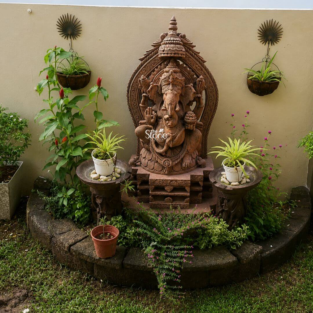 4.5 Feet Sandstone Ganesha Statue 3 A magnificent 4.5-foot Sandstone Ganesha statue serving as a prominent Ganesha stone Idol is elegantly positioned within a semicircular garden bed against a light-colored wall. Crafted from warm reddish-brown sandstone the statue depicts Lord Ganesha in a regal seated posture radiating benevolence and wisdom. His iconic elephant head is intricately carved featuring large ears expressive eyes and his distinctive broken tusk. He is adorned with an elaborate crown traditional jewelry and flowing garments all sculpted with remarkable detail emphasizing the natural texture and beauty of the sandstone. Ganesha is portrayed with four arms holding his customary attributes: a modak in one hand an axe or goad in another and his lower right hand extended in a gesture of blessing offering reassurance and protection. The statue is framed by a grand ornate arch or Prabhavali also carved from sandstone which features intricate traditional patterns floral motifs and perhaps mythical creatures further enhancing its divine presence and grandeur. The entire structure rests on a multi-tiered sandstone base elevating the deity within its garden sanctuary. The Ganesha idol is surrounded by a lush arrangement of green plants including ferns and smaller potted plants creating a harmonious and serene environment. Two smaller stone pedestals flank the main statue each topped with a white potted plant adding symmetry. Against the wall hanging planters with leafy green plants and hints of red flowers complement the scene. The semicircular stone border of the garden bed and the surrounding green grass suggest a thoughtfully designed outdoor space. This 4.5-foot Sandstone Ganesha statue is an exquisite example of a large outdoor deity designed to bring blessings peace and aesthetic grandeur to any garden or open-air setting perfectly aligning with an article focusing on such a significant Ganesha stone Idol.