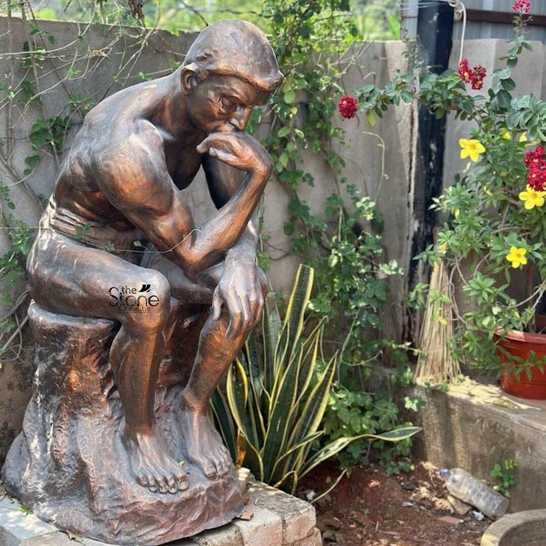 The Thinker Statue 3ft 3 A compelling 3-foot Fibre Thinking Man statue serving as an exceptional piece of Garden Decor is thoughtfully placed in a rustic outdoor setting surrounded by natural elements. The statue is an interpretation of Rodin's iconic "The Thinker depicting a muscular man seated in deep contemplation. His chin rests on his hand with his elbow on his knee and his brow is furrowed conveying intense intellectual engagement and introspection. The statue has a rich earthy bronze-like finish giving it a classic and artistic appeal with visible textures that mimic cast metal. The figure's powerful physique is sculpted with strong defined lines highlighting the tension and depth of thought. He is depicted in a mostly nude state with a simple drapery around his waist emphasizing the human form and its capacity for profound thought. The statue is seated on a rough organic pedestal that resembles a tree stump or a natural rock formation further integrating it into the garden environment. This choice of material and finish makes it durable and aesthetically suitable for outdoor display. The statue is positioned on a paved or stone area with a bed of soil and various green plants including a variegated snake plant in the foreground creating a natural and contemplative atmosphere. In the background climbing vines cover a wall and flowering plants with yellow and red blooms add splashes of color and vitality. This 3-foot Fibre Thinking Man statue is an exquisite example of garden art designed to inspire contemplation intellectual curiosity and aesthetic beauty in any outdoor space perfectly aligning with an article focusing on such a profound and artistic Garden Decor Thinking Man Statue.