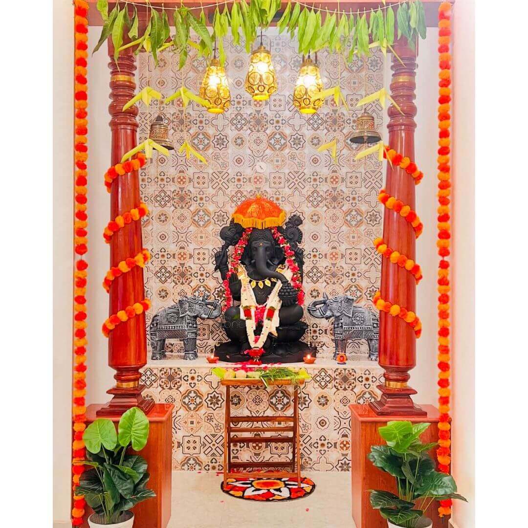 Fiber Ganesha Statue 3.5ft 8 beautifully decorated home temple or puja altar, radiating a vibrant and spiritual ambiance. At the center, a majestic black stone idol of Lord Ganesha is seated, adorned with a prominent white and red floral garland, possibly of jasmine and rose petals, draped over his chest. A smaller red garland also graces his form, and a bright orange ceremonial umbrella (chhatri) is placed above his head, signifying respect and divinity. Ganesha is flanked on either side by two intricately carved grey elephant statues, detailed with traditional patterns, adding to the regal and auspicious atmosphere. This central arrangement rests on a raised platform, where several small lit diyas (oil lamps) cast a warm glow. The backdrop to this sacred display is a wall covered in ornate, earthy-toned tiles featuring intricate geometric and floral motifs in shades of brown, beige, and white. Framing the entire altar are two substantial, dark reddish-brown wooden pillars with traditional Indian carvings, creating a classic temple-like structure. These pillars are elegantly wrapped with long garlands of bright orange and yellow marigold flowers, which also hang vertically along their sides, adding bursts of color. Above the altar, a canopy of fresh green mango leaves is strung horizontally, forming a festive arch, a common decoration for auspicious occasions. From this overhead structure, three ornate, warm-toned hanging lamps, possibly made of brass or intricately patterned glass, cast a soft, inviting light. Small golden bells are also tied to the pillars near the top, hinting at a gentle chime. In front of Ganesha, on the altar, a small wooden stand with shelves holds various ritualistic items, including green leaves, likely for offerings. On the floor directly below, a colorful circular rug with a vibrant floral pattern in shades of red, orange, black, and white adds a touch of warmth and design. At the base of each wooden pillar, large potted green plants with broad, glossy leaves in simple white planters bring a natural, refreshing element to the scene. The entire setup perfectly illustrates a traditional Hindu spiritual space, ready for daily puja or festive celebrations like Ganesh Chaturthi, embodying devotion, rich cultural aesthetics, and the symbolism associated with welcoming prosperity and removing obstacles.