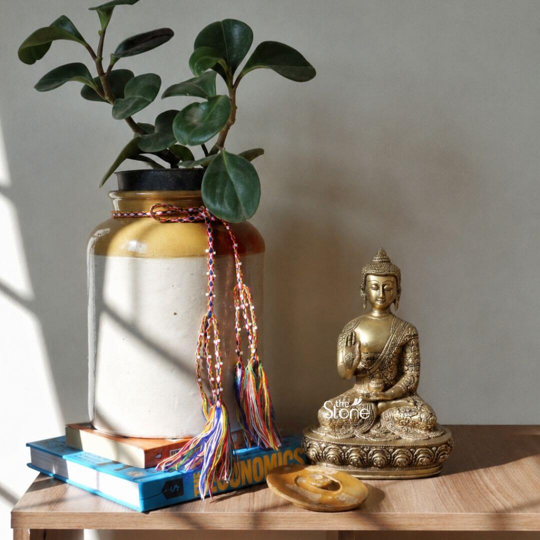Small Brass Buddha Statue 10" 3 A small 10-inch brass Buddha statue is displayed on a wooden tabletop as part of a warm and thoughtfully arranged home décor setting. The Buddha is seated cross-legged on an ornate lotus-style pedestal with one hand raised in a blessing or reassurance gesture and the other resting gently in the lap. The figure has a calm facial expression softly lowered eyes elongated earlobes detailed curled hair and a pointed ushnisha while the richly patterned robe and finely carved jewellery-like motifs highlight the craftsmanship of the brass sculpture. The golden-toned Buddha statue is positioned beside a large cream and mustard ceramic planter holding a healthy green indoor plant with broad glossy leaves. The planter rests on a small stack of books and is decorated with colourful braided cords and hanging tassels adding a relaxed artistic character to the arrangement. A smooth natural stone object lies in front of the statue while sunlight enters from the left and casts soft geometric shadows across the wall planter books and wooden surface creating a peaceful and inviting atmosphere. This image demonstrates how a Small Brass Buddha for Home Decor can be styled as an elegant spiritual accent on a console table bookshelf study desk, sideboard or meditation corner. The compact 10-inch Brass Buddha statue offers enough visual presence to become a meaningful focal point without overwhelming the surrounding décor. Its antique brass finish detailed lotus base serene posture and placement among books, greenery natural textures and warm sunlight convey mindfulness harmony and timeless beauty within a contemporary home interior.
