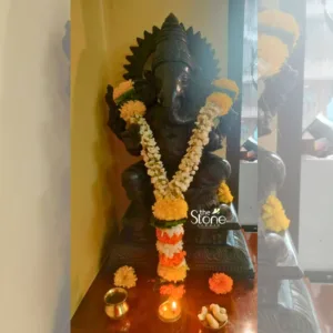 majestic, dark stone statue of Lord Ganesha, crafted from what appears to be dark, polished granite or a similar heavy stone, stands prominently on a polished dark brown wooden altar or pedestal. The deity is depicted in a traditional seated posture, featuring an elephant head with large, intricately detailed ears, a gracefully curved trunk, and four arms, holding various auspicious symbols (though specific details of the items in hand are obscured by decorations). Ganesha is adorned with a detailed crown and a radiant, spoked halo (prabhāmaṇḍala) behind its head, indicating its divine presence. The Ganesha idol is richly decorated with fresh floral garlands. A long, U-shaped garland of delicate white jasmine flowers, interspersed with vibrant yellow marigold pom-poms, drapes elegantly around its neck and chest. Below this, a shorter, tiered garland of bright orange and yellow marigold flowers, possibly accented with subtle green leaves, adds another layer of festive color. Several loose orange and yellow marigold blooms are scattered artfully around the base of the statue, enhancing the devotional atmosphere. On the polished dark brown wooden surface directly in front of the statue, several traditional puja offerings are arranged: a small, lit brass diya (oil lamp) casting a warm, flickering glow; a gleaming brass pot or kumkum holder; and a small brass plate filled with white cashew nuts, representing prasad (sacred offerings). The setting features a plain, warm-toned beige wall to the left, and a softly blurred, lighter background on the right, possibly a window or a reflective surface, which adds depth and helps focus attention on the central figure. The overall image evokes a sense of sacred ritual, devotion, and tranquility, showcasing exquisite craftsmanship. The "the Stone STUDIO" logo is visible in the lower right, suggesting this beautiful Ganesha idol is a high-quality work of art or a product from a specialized studio dealing in traditional stone carvings, religious sculptures, deity statues, or handcrafted spiritual decor for home altars and temples. This detailed visual representation highlights traditional Indian religious art, puja arrangements, and cultural aesthetic, making it highly relevant for articles discussing Hindu deities, devotional practices, spiritual home decor, or the artistry of stone carving.