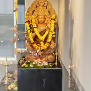 magnificent, richly detailed stone sculpture of the Hindu goddess Saraswati, carved from a warm, golden-brown stone, possibly sandstone or a similar material, stands as the central focus of the image. She is depicted in a serene, seated posture, likely Lalitasana (one leg folded, the other gently hanging), upon an elaborately carved lotus pedestal. Her four arms gracefully hold her characteristic attributes: the veena (a stringed musical instrument) is held across her lap, while her upper right hand holds a rosary (mala), and her upper left hand a scripture (pustaka), symbolizing knowledge and arts. Saraswati wears a majestic crown and is adorned with finely sculpted jewelry, including necklaces, armlets, and anklets, emphasizing her divine form. Behind her head, an ornate Prabhavali (aura or arch) frames her, featuring traditional Hindu motifs and intricate scrollwork, enhancing her divine presence and craftsmanship. The sculpture is ceremonially adorned with two thick, vibrant garlands of bright yellow and orange marigolds, draped around her neck and shoulders, falling in a V-shape down to her lap, adding a celebratory and devotional touch. At the base of the statue, on a dark, polished stone pedestal, are ceremonial offerings. These include fresh green banana leaves laden with a colorful array of flowers (white jasmine, pink roses, red hibiscus petals) and several green bananas, typical of Hindu puja rituals. Multiple gleaming brass oil lamps (diyas) with flickering flames are placed symmetrically around the offerings and extend along the perimeter of the pedestal, casting a warm, inviting glow. The entire setup is against a simple, light-colored wall, creating a clean backdrop that allows the intricate details of the sculpture and its adornments to stand out. Reflections on the left side of the image suggest a window or light source nearby. A discreet logo, "the Stone STUDIO," is visible in the lower left corner of the statue, indicating the craftsmanship behind this stunning piece. This image effectively showcases the artistry of stone carving, traditional Hindu deity sculptures, and the reverence associated with such installations, making it ideal for an article discussing custom stone craftsmanship, spiritual art, or traditional Indian temple sculpture.